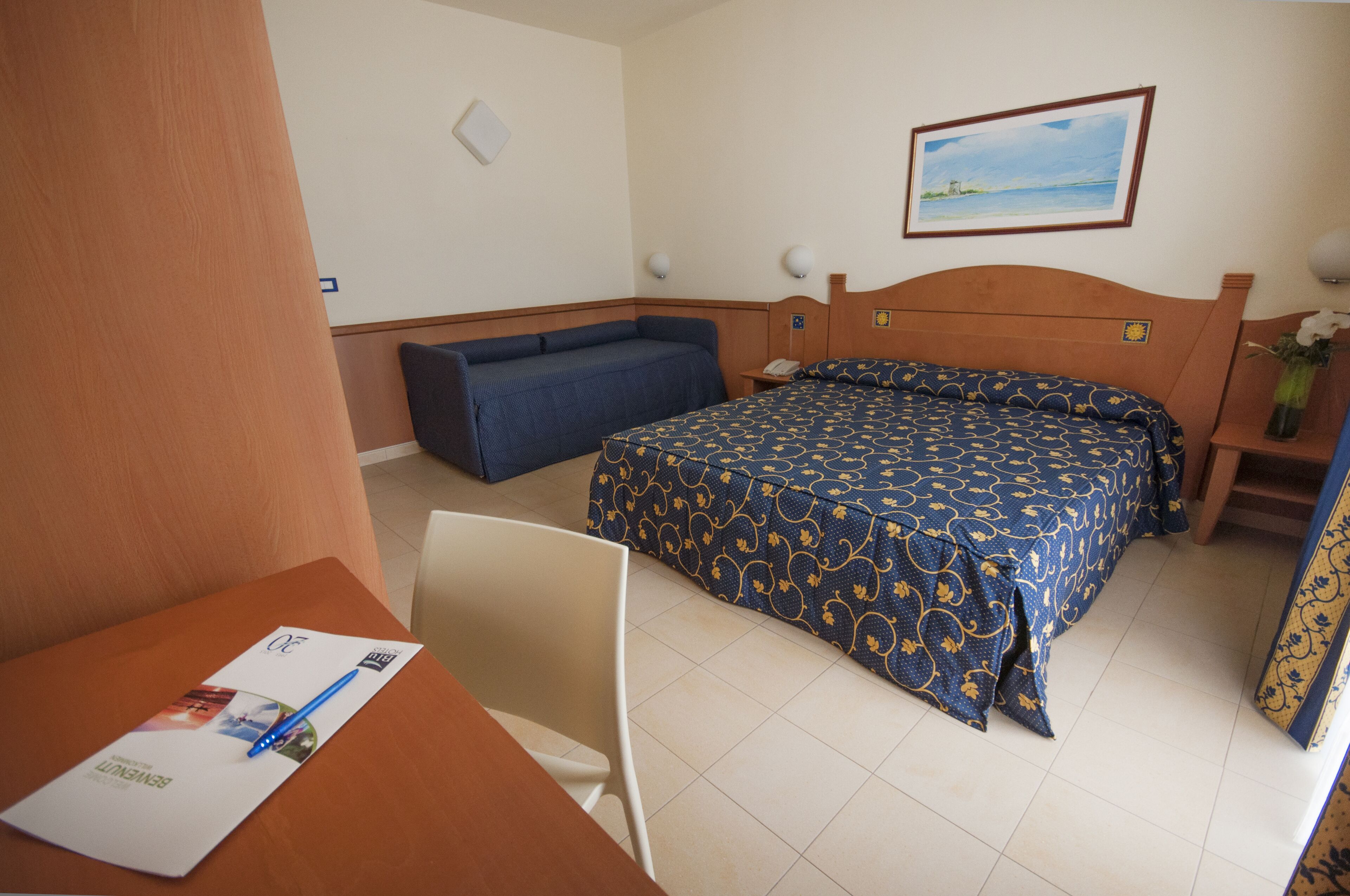 A hotel room with a bed, a desk, and a chair. There is a picture on the wall and a vase with flowers on the nightstand.