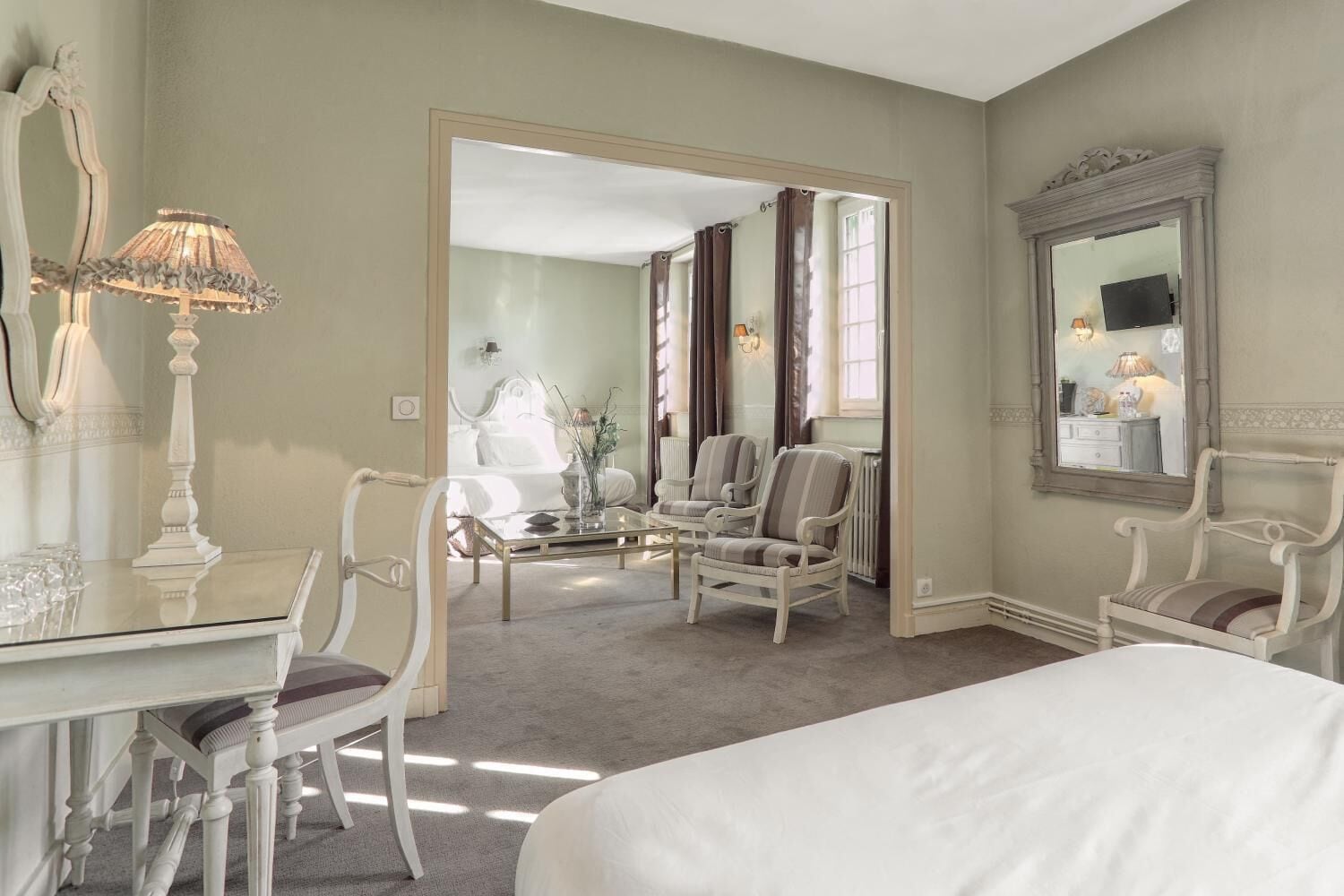 Une chambre d’hôtel avec un grand lit, un coin salon avec des fauteuils, une table avec des verres et un miroir au mur.