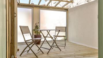 An open patio with a wooden table and two chairs, a potted plant, and a view through a glass door.