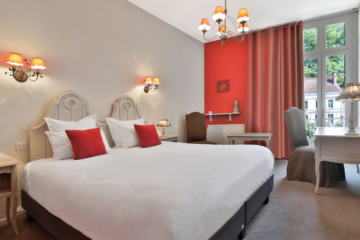 Une chambre d’hôtel avec un grand lit, deux tables de chevet, un bureau et une chaise. Il y a une fenêtre avec des rideaux, un lustre et des appliques murales.