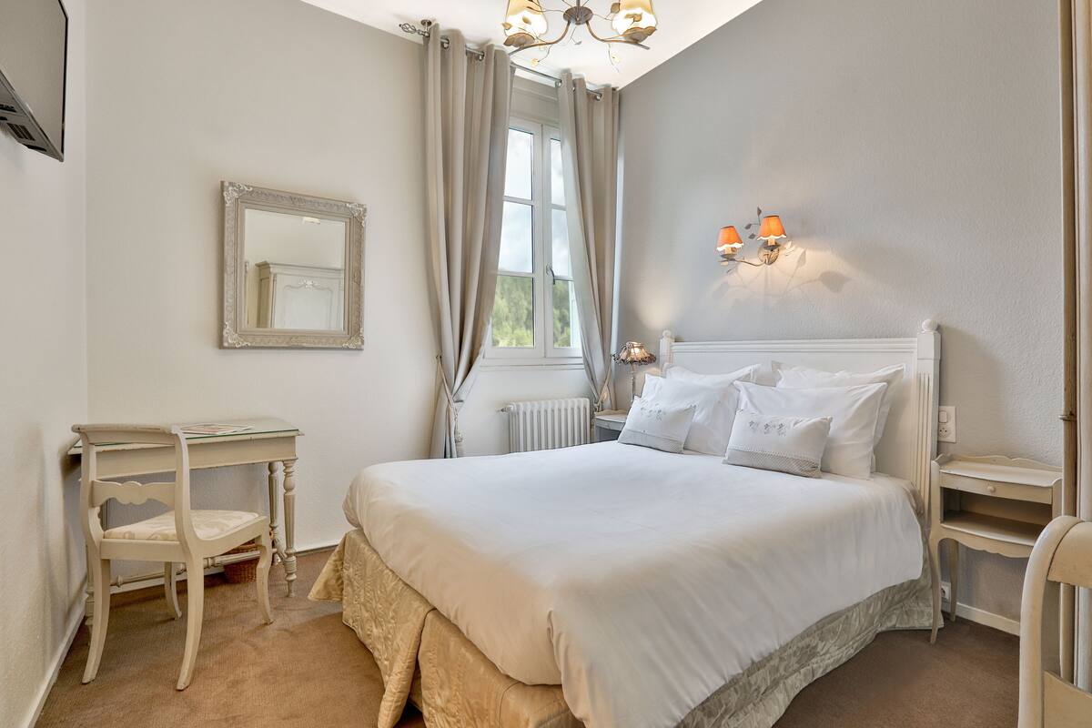 Une chambre avec un grand lit, une table de chevet, une chaise, un miroir et une fenêtre avec des rideaux.