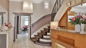 A hotel reception area with a wooden counter, a staircase with a decorative railing, and a mirror above the counter.