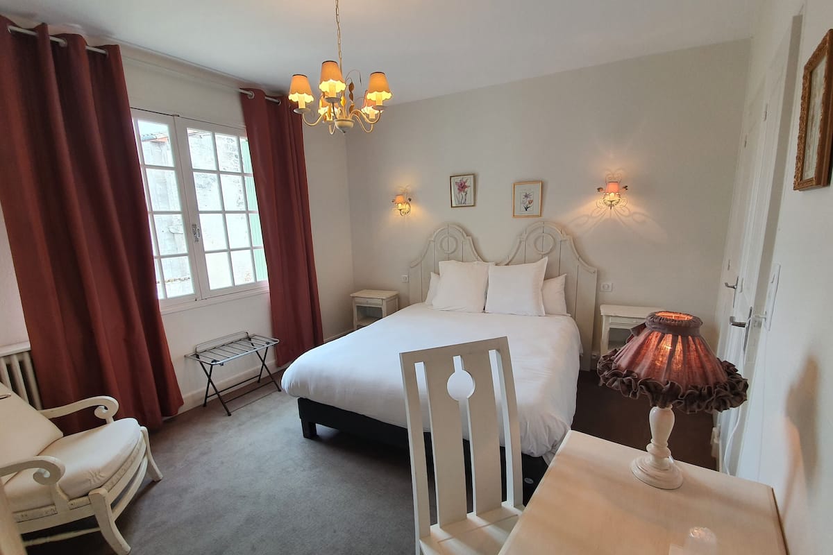 Une chambre à coucher comprenant un lit, une chaise, une table, un luminaire suspendu et une fenêtre avec des rideaux.