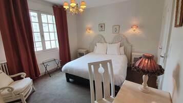 A bedroom with a bed, a chair, a table, a hanging light fixture, and a window with curtains.