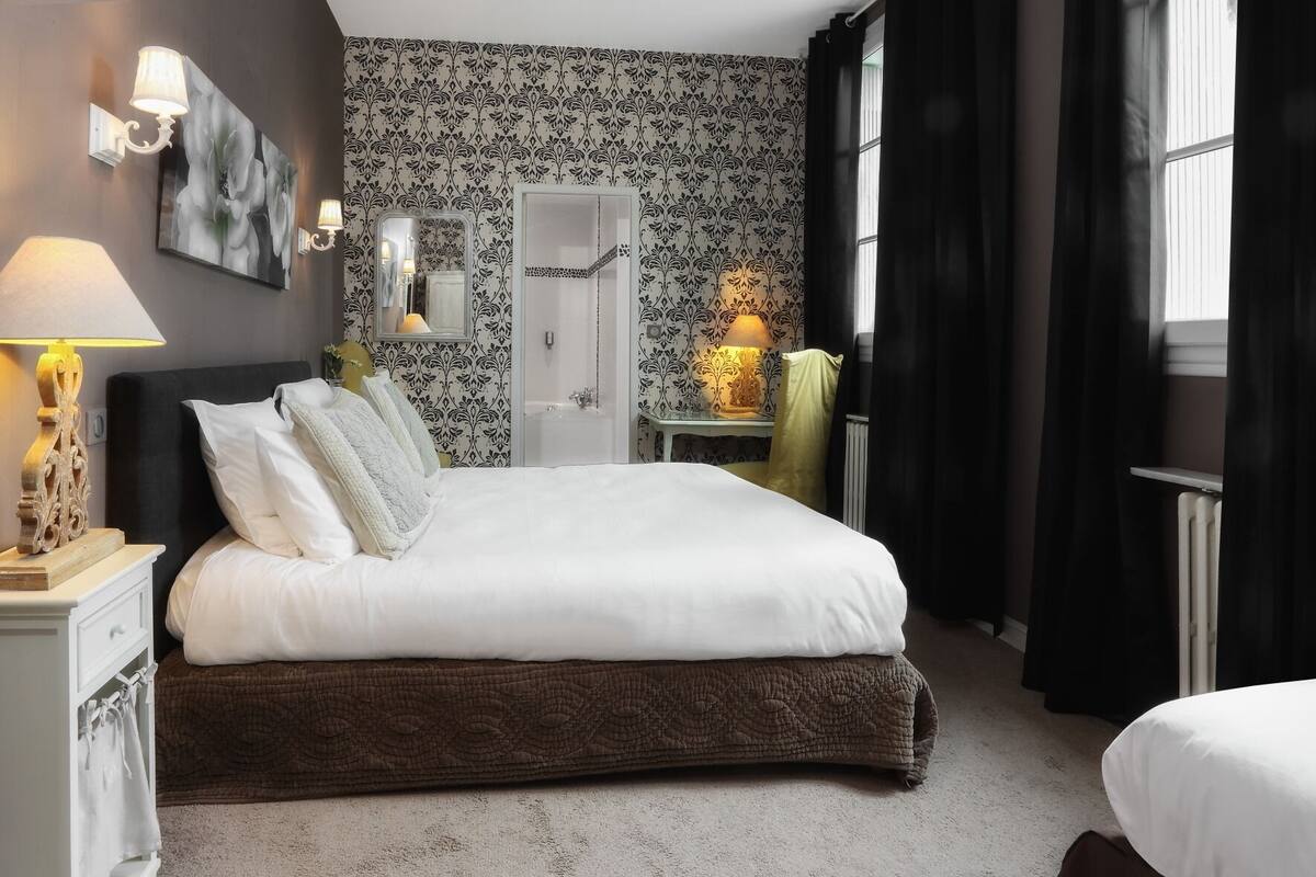 Une chambre à coucher avec un grand lit, une table de chevet avec une lampe, un miroir, une commode et une fenêtre avec des rideaux.