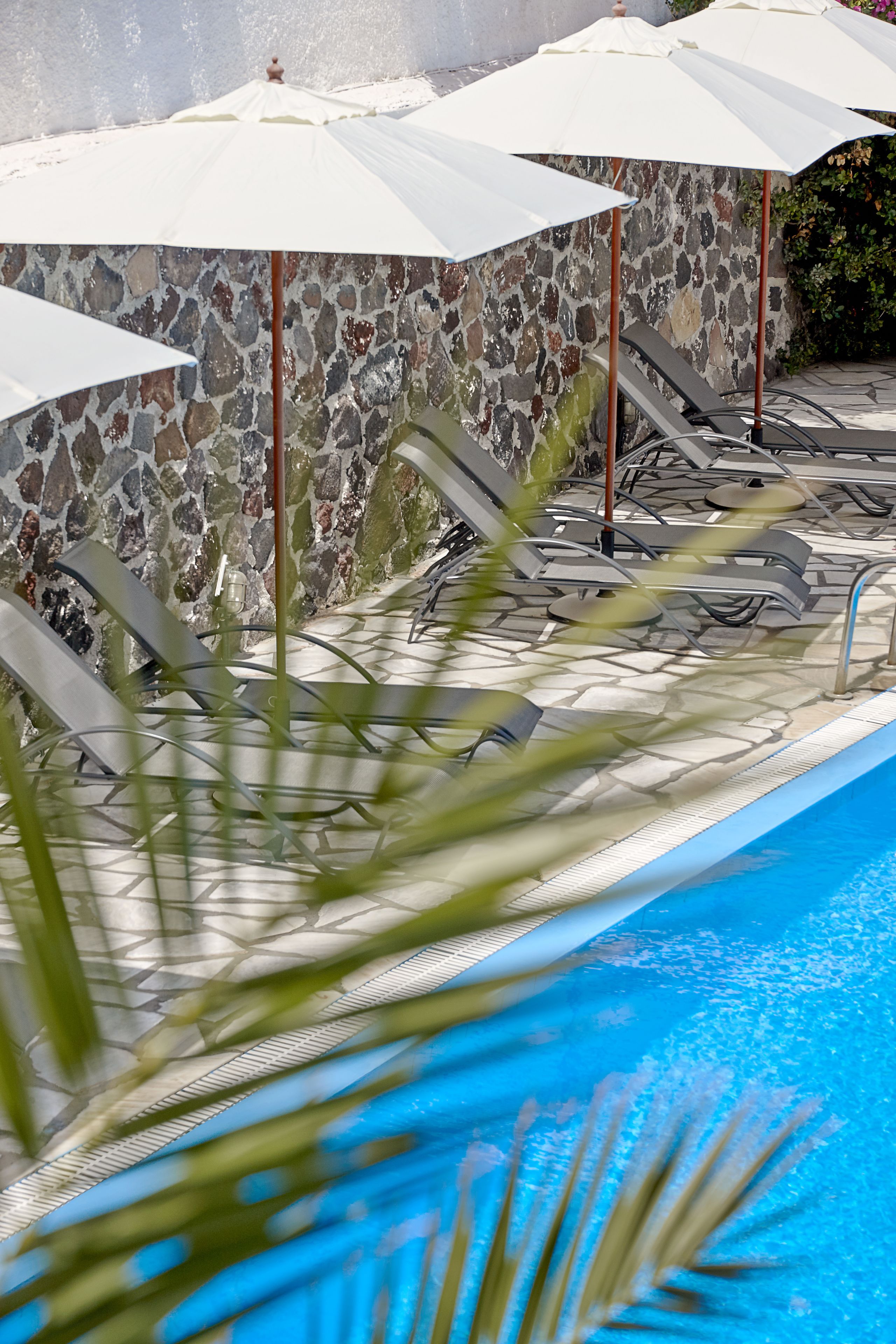 A pool area with lounge chairs and umbrellas, a stone wall, and a clear blue pool.