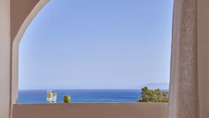 A view through an arched window overlooking a body of water and distant land, with a glass of water and an apple on the ledge. - Terra Blue Santorini (Santorini)