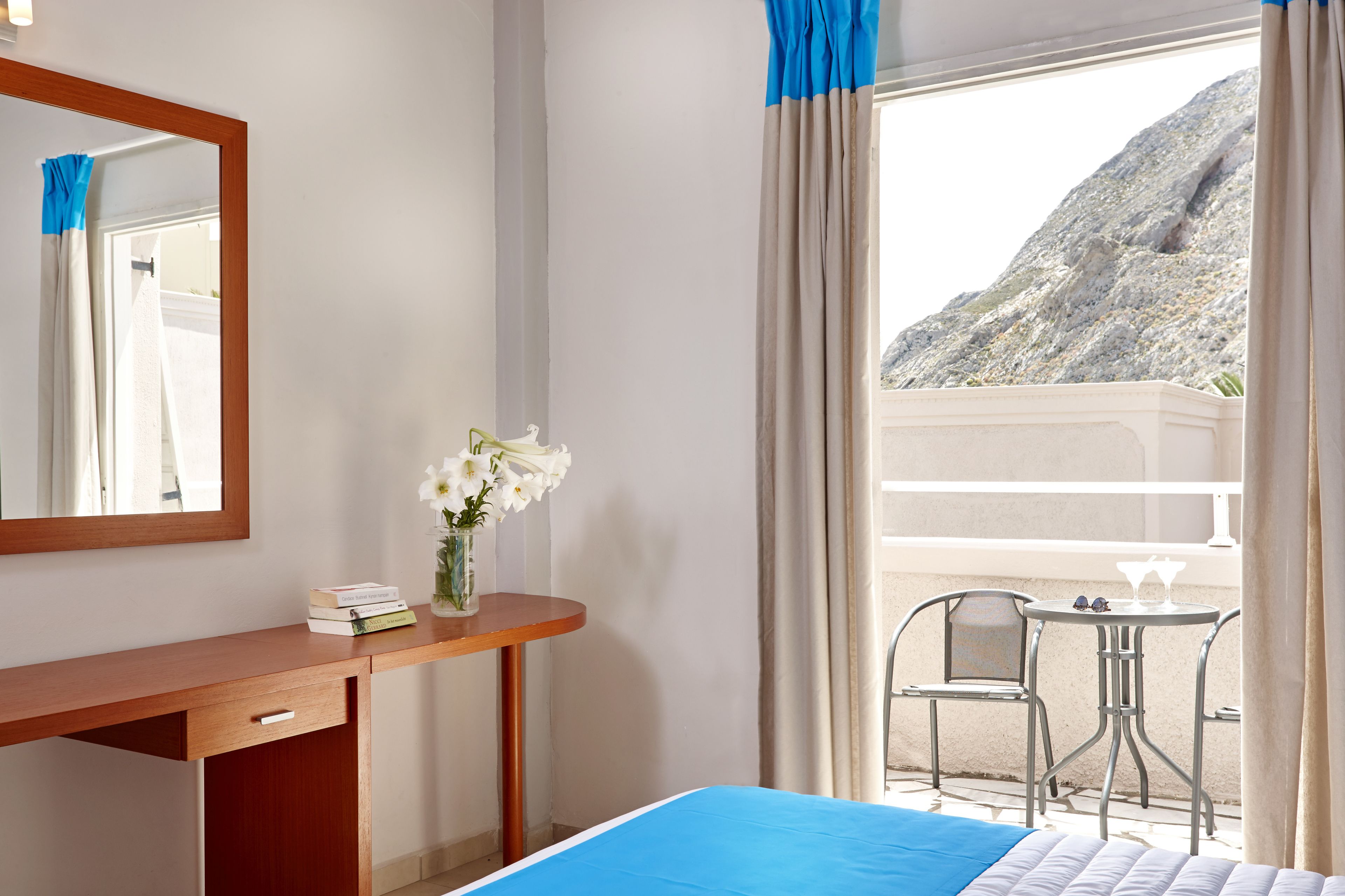 A hotel room with a bed, a desk with a mirror, a vase with flowers, and a balcony with a table and chairs.