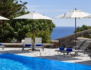 A poolside area with lounge chairs and umbrellas, a clear view of the ocean, and a stone wall. - Terra Blue Santorini (Santorini)