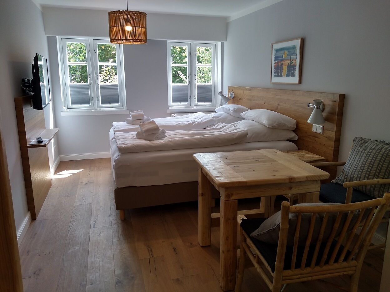 A bedroom with a bed, a wooden table, and a chair. There are windows with blinds, a hanging light fixture, and a painting on the wall.