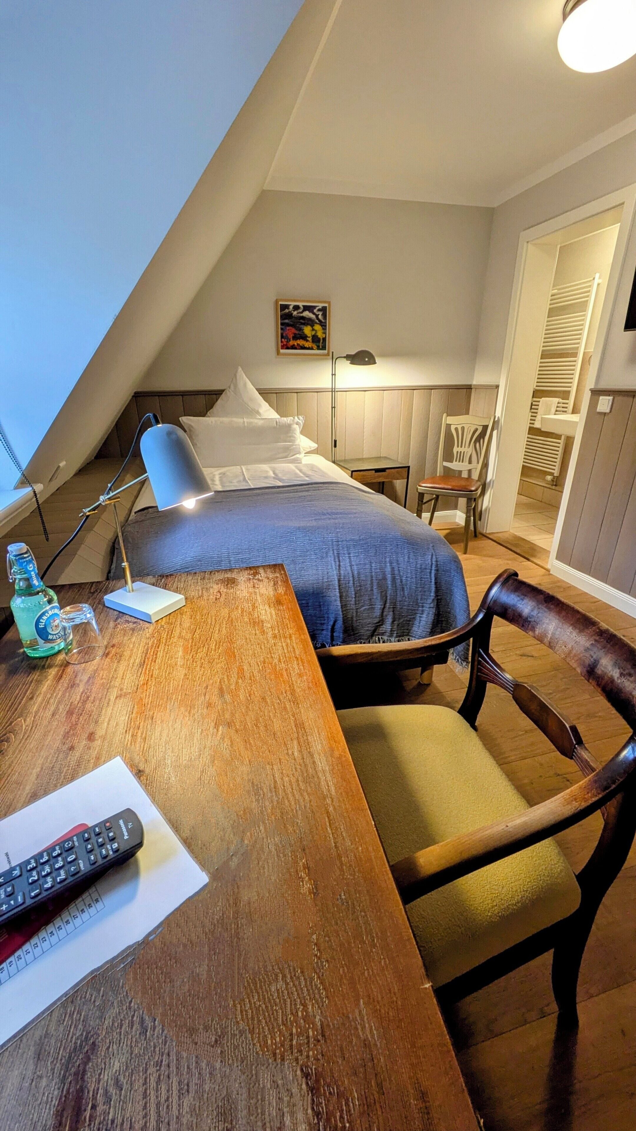 A bedroom with a bed, a desk with a lamp, a chair, a remote control, and a bottle of water.