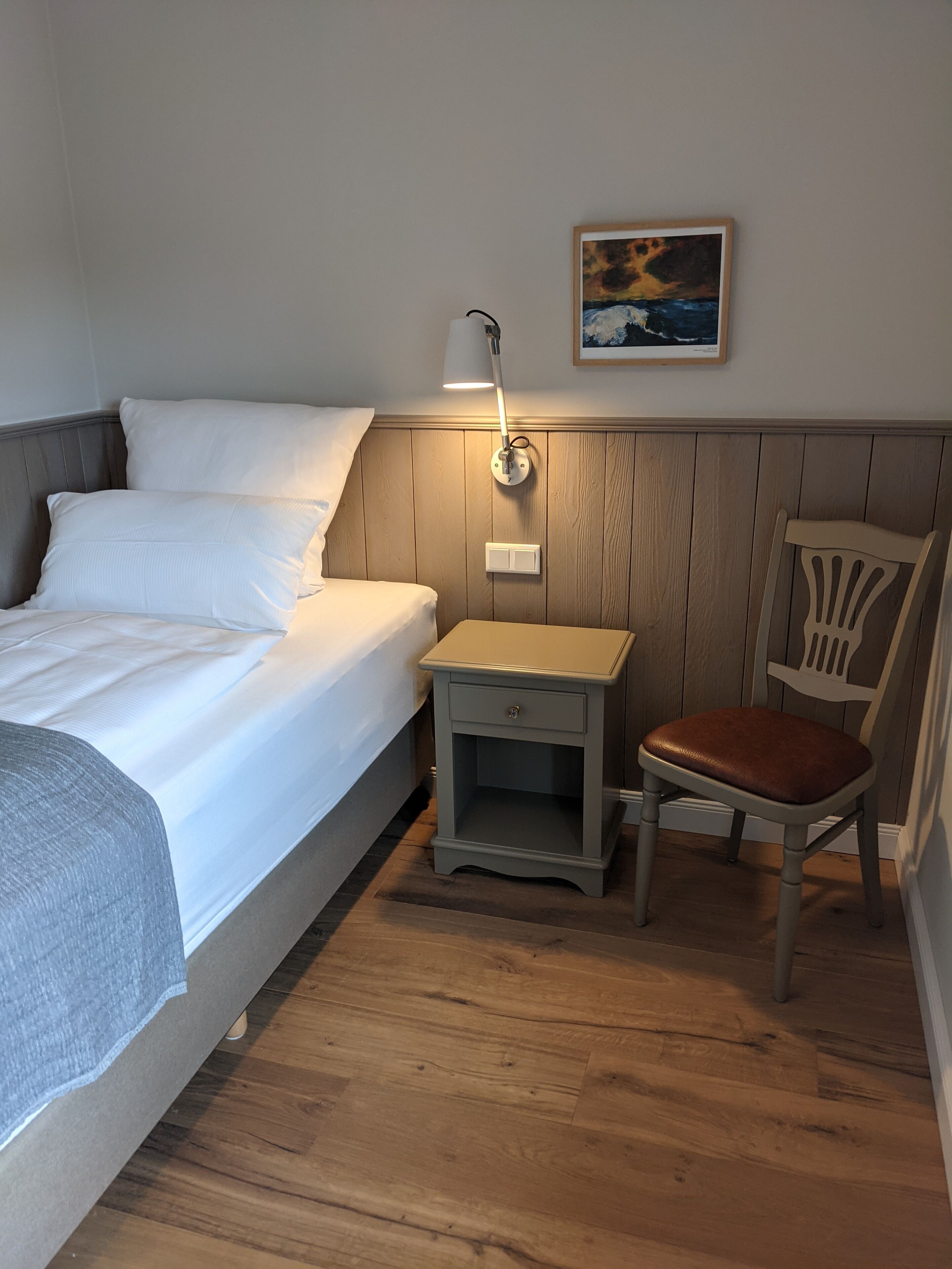 Un dormitorio con cama, mesita de noche, silla y un cuadro en la pared.