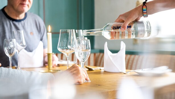 Une personne verse de l’eau dans un verre sur une table où se trouvent une bougie et d’autres verres.