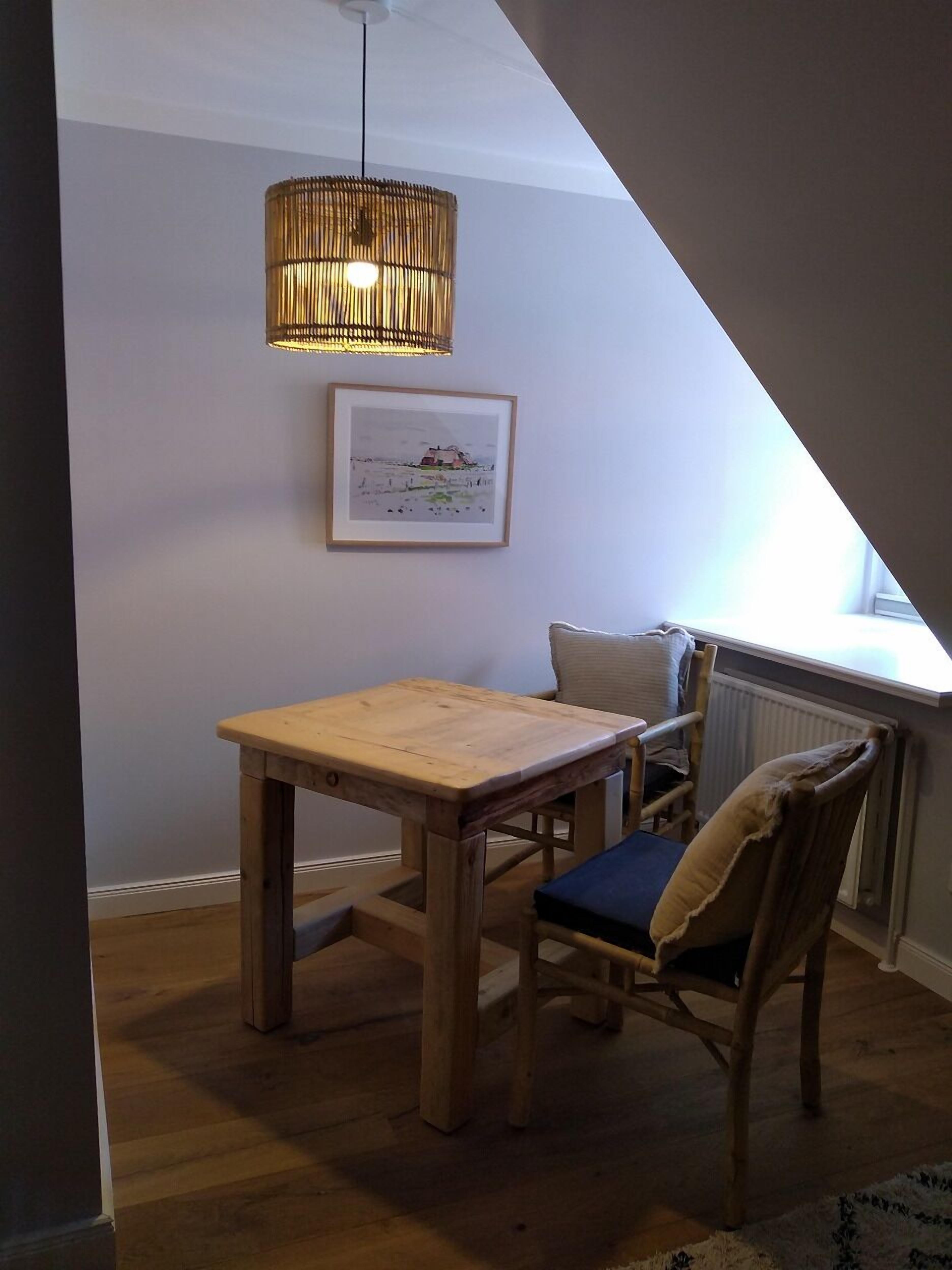 A loft-style room with a wooden table and chair, a hanging light fixture, a framed picture on the wall, and a window with a view.