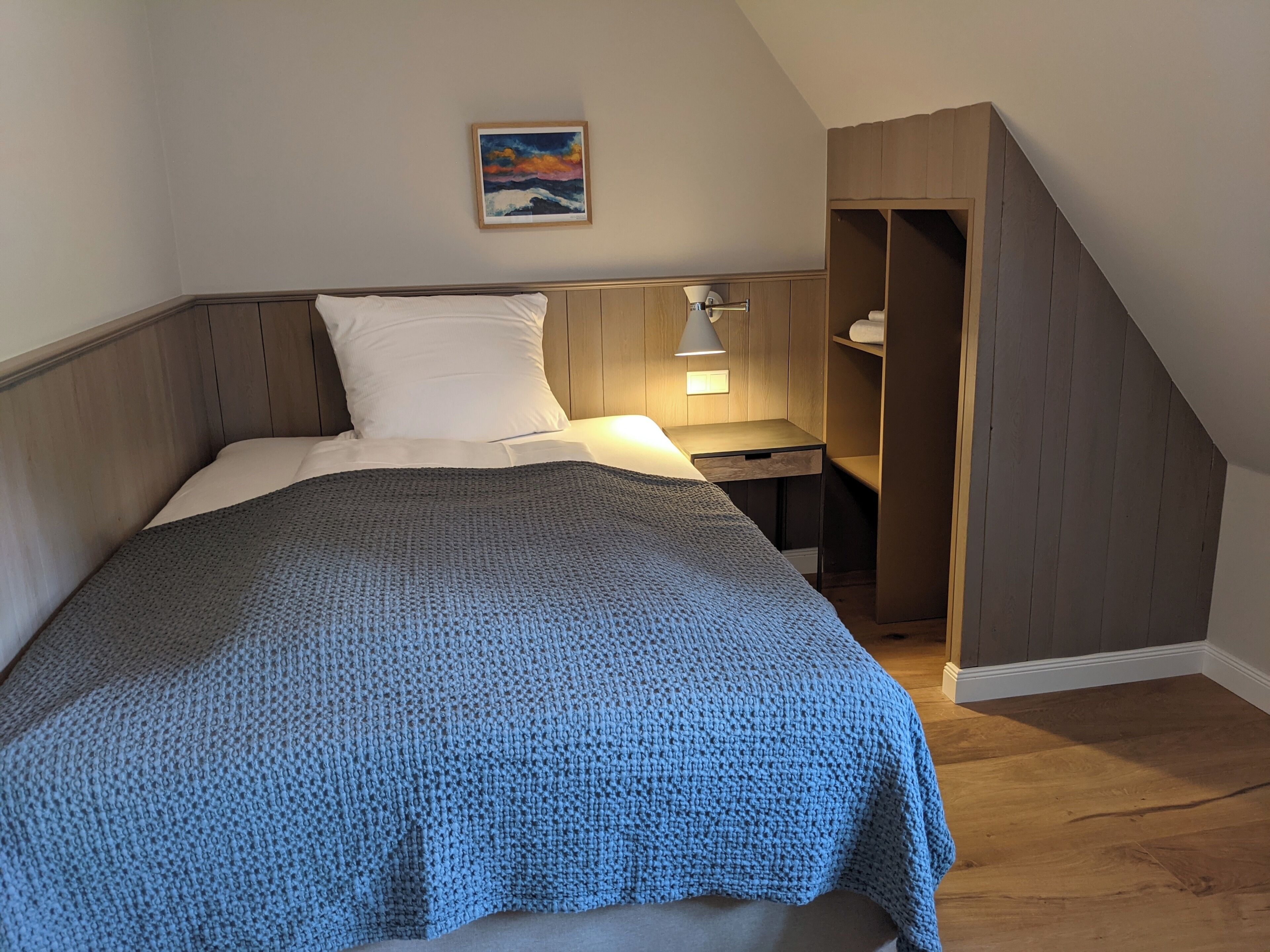 A bedroom with a bed, a nightstand, a lamp, a painting on the wall, and a wooden wardrobe.
