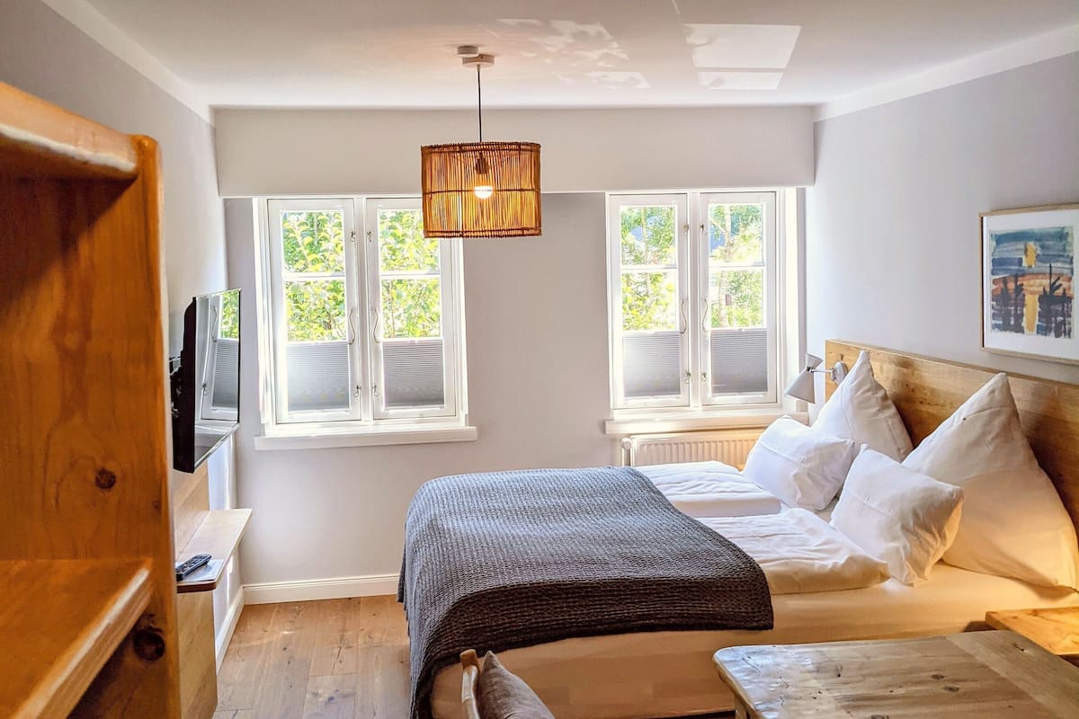 Une chambre à coucher de taille réduite, comprenant un lit, des tables de chevet, un banc et un luminaire suspendu.