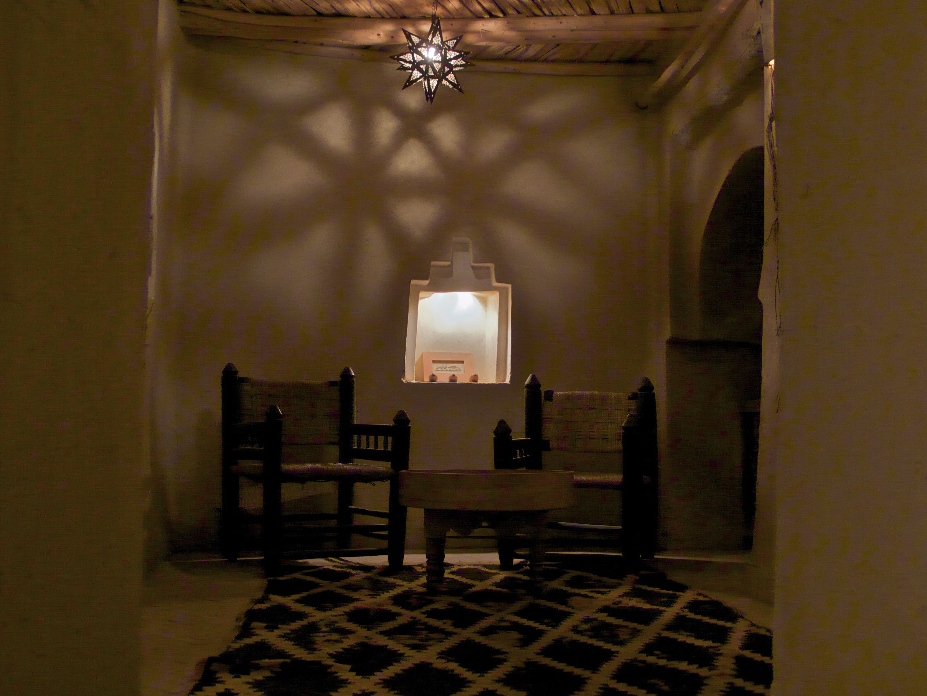 Um quarto com iluminação suave, um tapete estampado, uma cadeira de madeira, uma mesa pequena e um lustre decorativo em formato de estrela sobre uma janela.