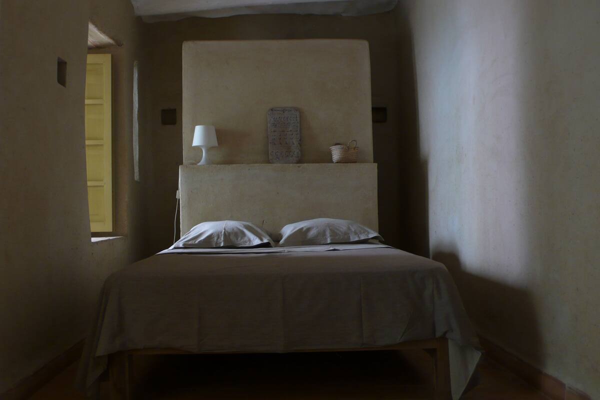 A small, dimly lit room with a single bed, a nightstand, a lamp, and a wall-mounted shelf with a basket.