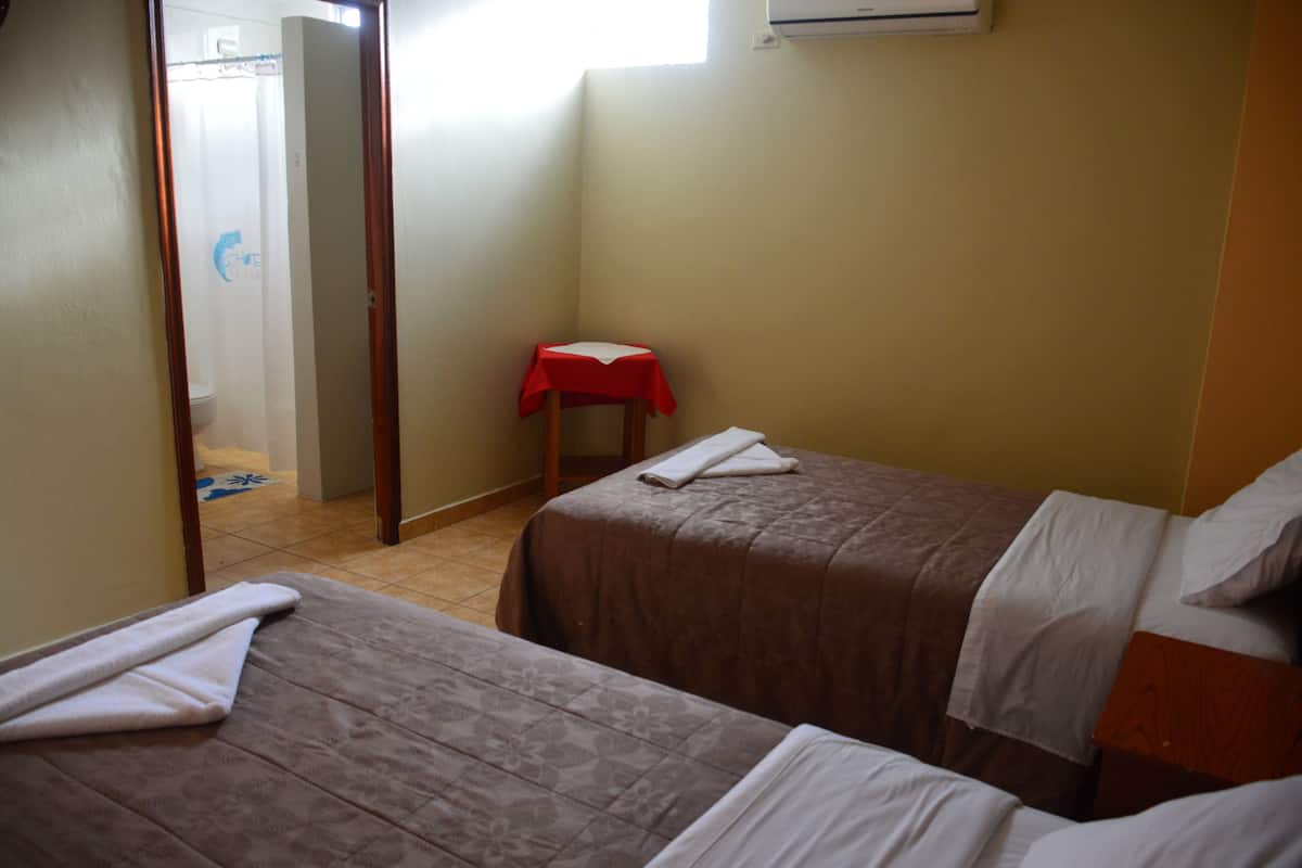 A hotel room with two beds, a small table with a red tablecloth, and a shower area.