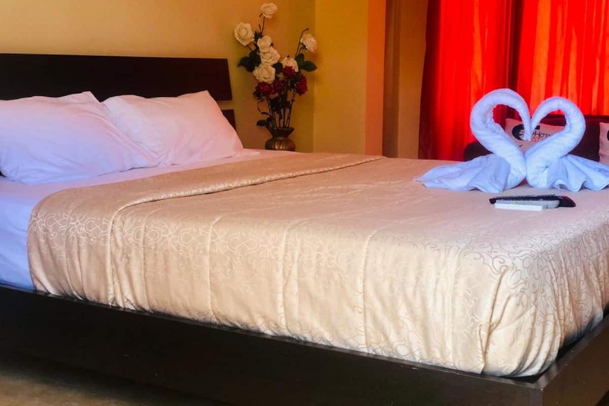 A neatly made bed with a beige bedspread and pillows, a vase of white flowers, and a heart-shaped towel arrangement on the bed.