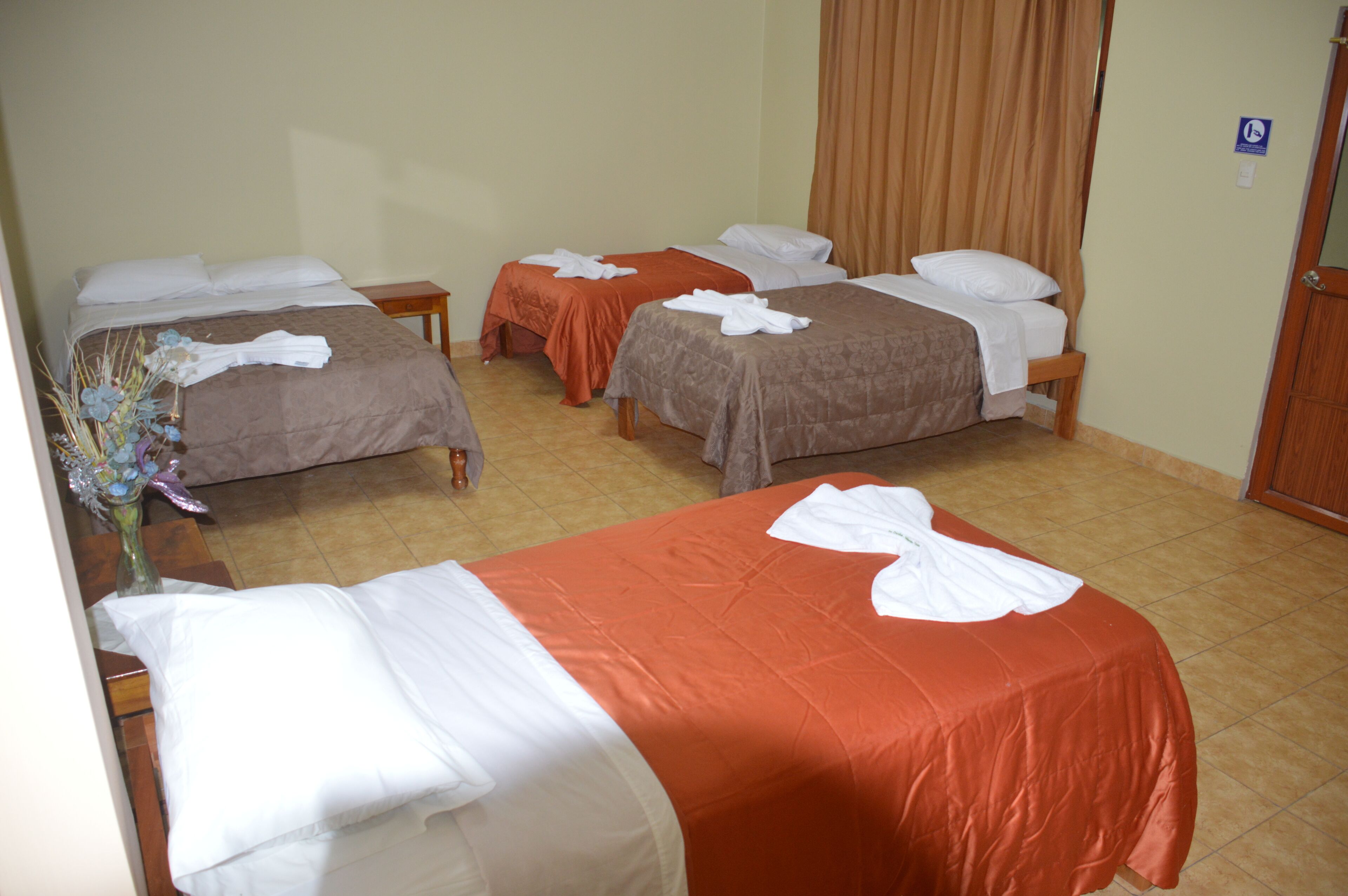 A hotel room with three beds, each with a white towel and brown bedsheets. There is a wooden nightstand with a vase of flowers.