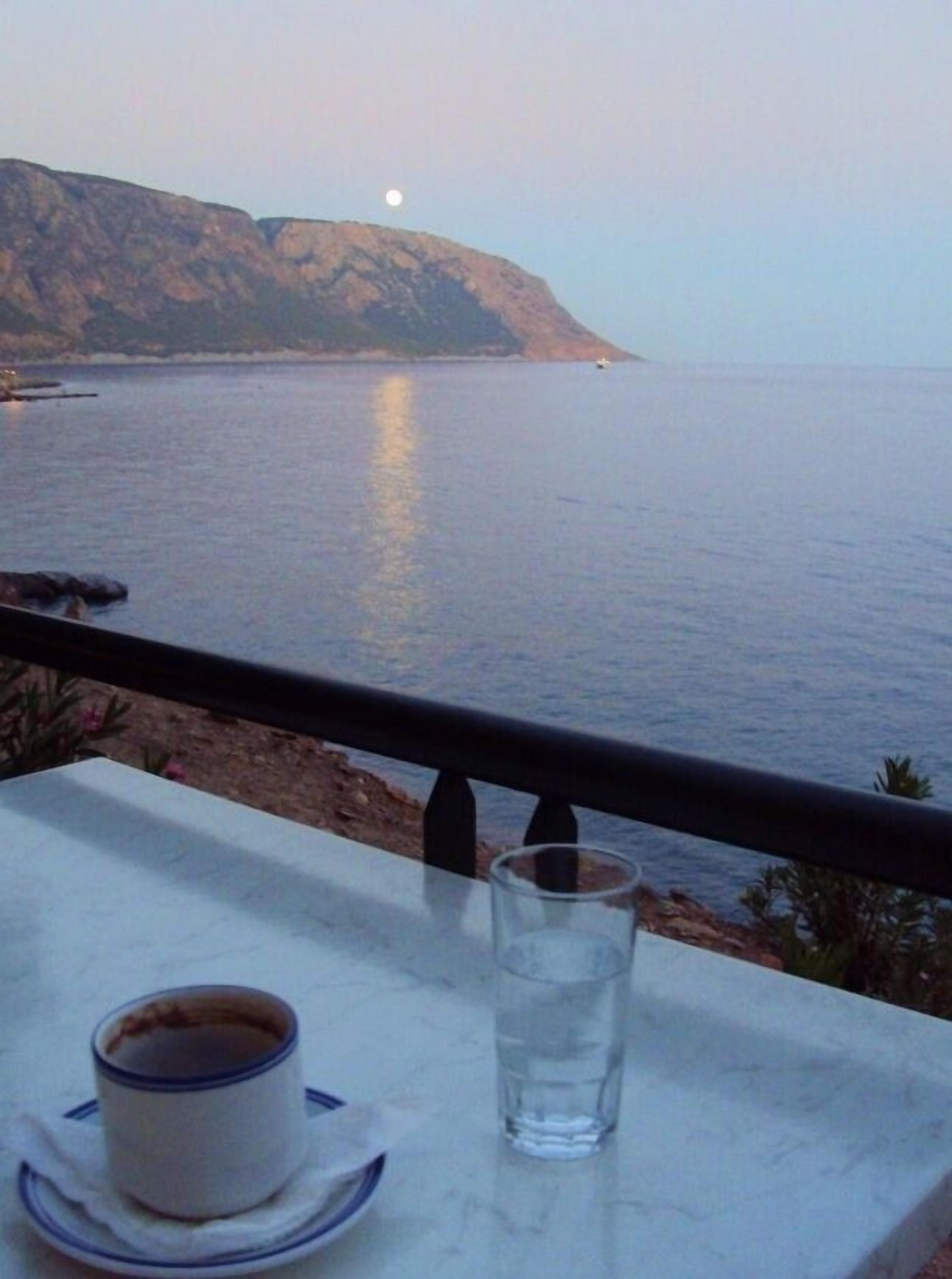 Ein Tisch mit Tasse und Glas, mit Blick auf ein ruhiges Meer und einen fernen Berg.