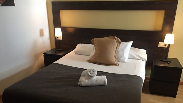 A neatly made bed with a dark bedspread, two pillows, and a rolled towel. There are two bedside tables with lamps. - Sitges Go (Sitges)