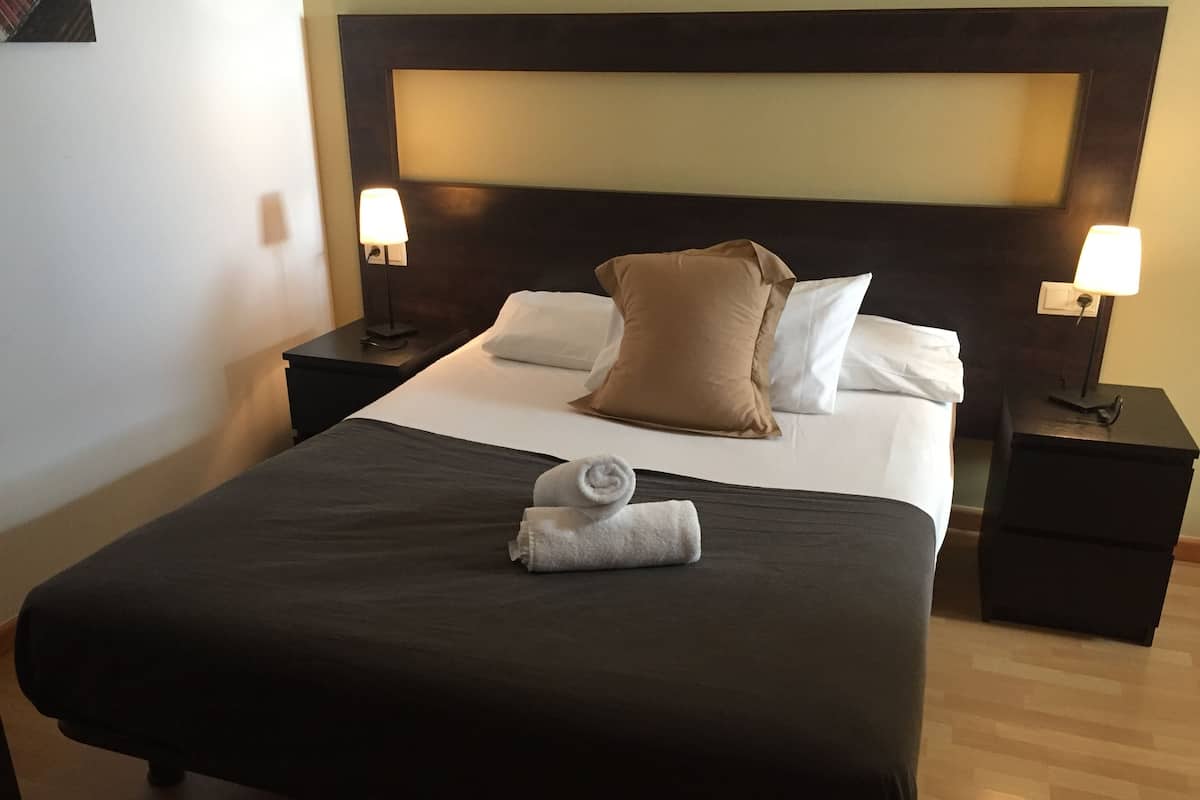 Ein ordentlich gemachtes Bett mit dunkler Tagesdecke, zwei Kissen und einem zusammengerollten Handtuch. Es gibt zwei Nachttische mit Lampen.