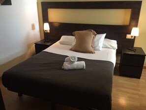 Ein ordentlich gemachtes Bett mit dunkler Tagesdecke, zwei Kissen und einem zusammengerollten Handtuch. Es gibt zwei Nachttische mit Lampen.