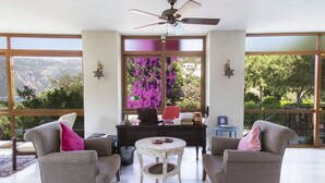 Una habitación con dos sillones grises, una mesita redonda pequeña, un ventilador de techo y un escritorio con una computadora portátil y objetos decorativos. Amplias ventanas ofrecen vistas a árboles y montañas.