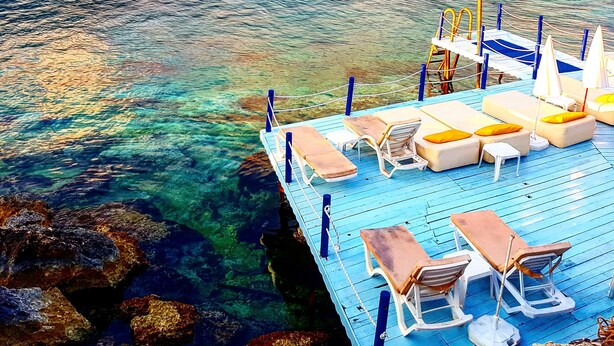 Un muelle con agua cristalina que lleva a un paisaje montañoso. Hay sillas de descanso y sombrillas en el muelle.