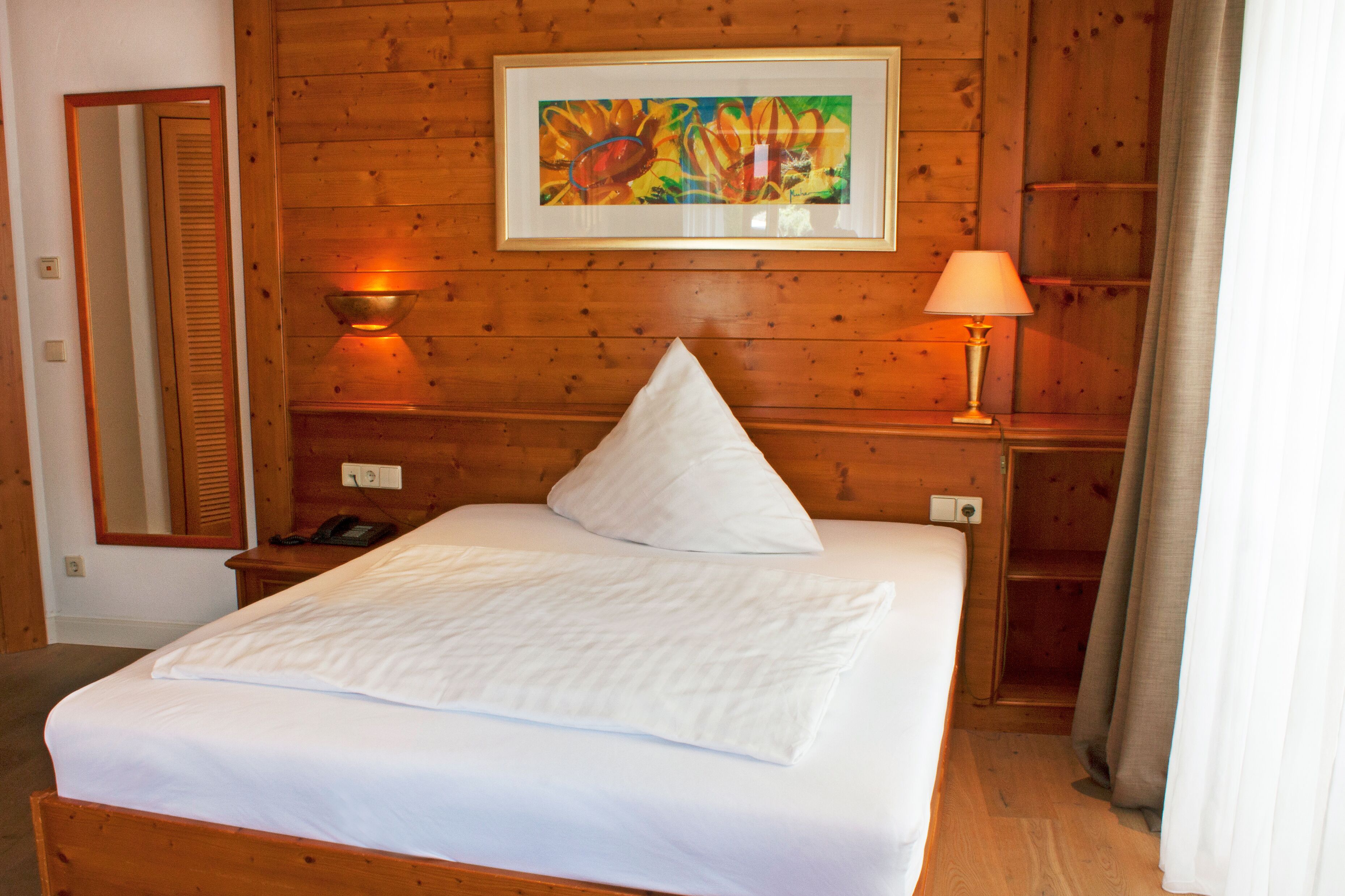 A wooden-paneled room with a bed, a bedside lamp, a telephone, and a framed painting on the wall.