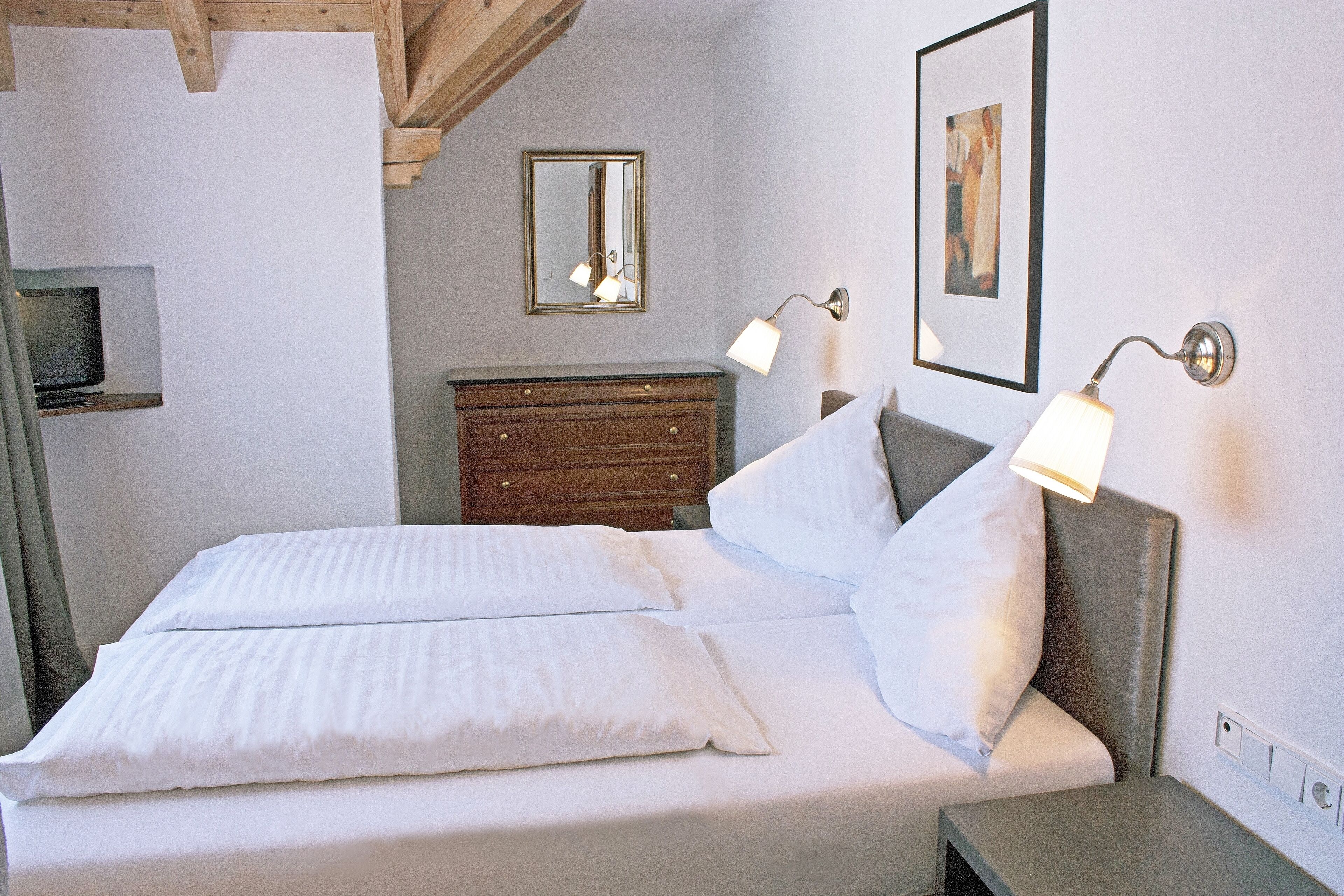 Una cama bien hecha con ropa blanca, un aparador de madera, un espejo y dos lámparas fijadas a la pared.