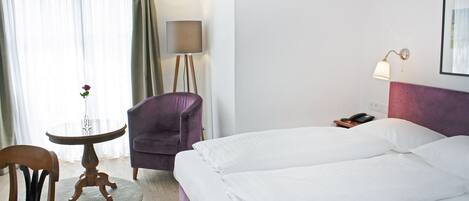 Habitación de hotel con una cama, una silla, una mesita con un florero, una lámpara y un teléfono en una mesita de noche.