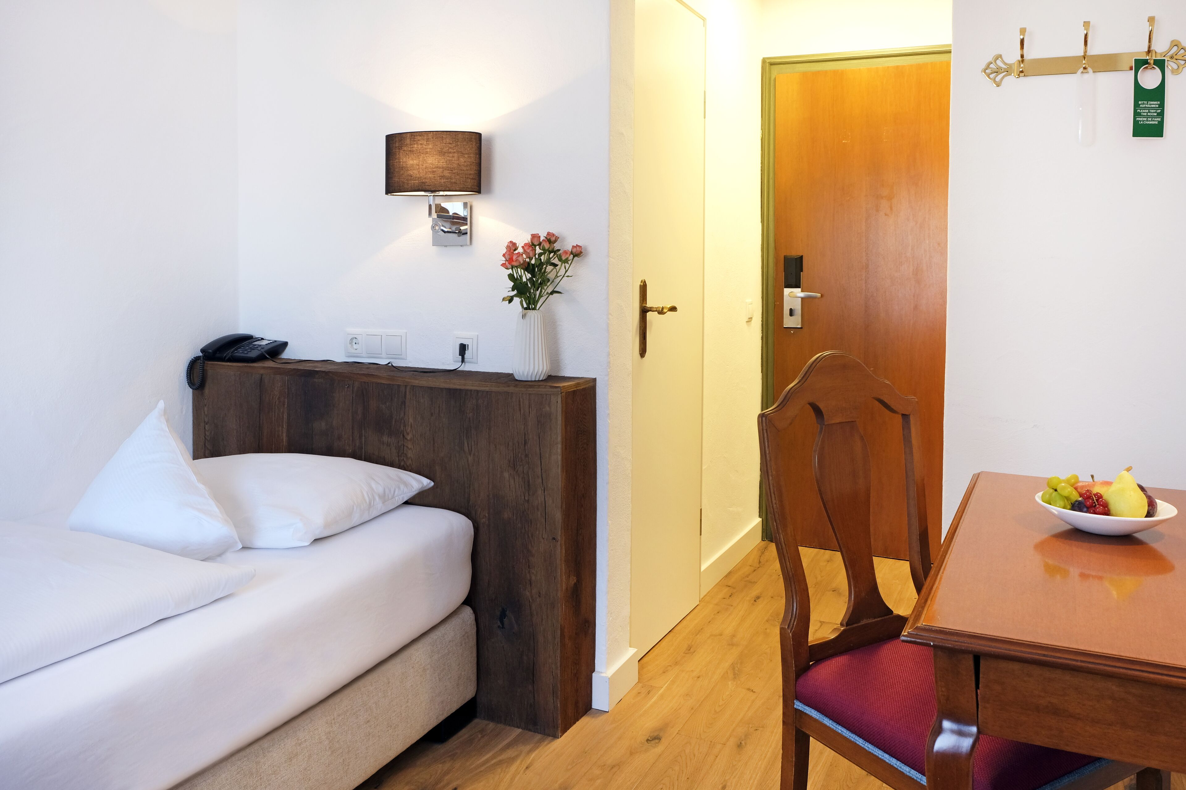 Ein Hotelzimmer mit Bett, Nachttisch, Lampe, Blumen in einer Vase, Holztür und einem Esstisch mit Obst.