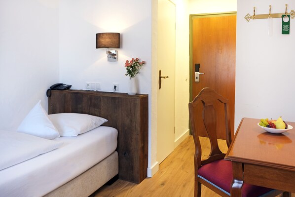 A hotel room with a bed, bedside table, lamp, vase with flowers, wooden door, and a dining table with fruit.