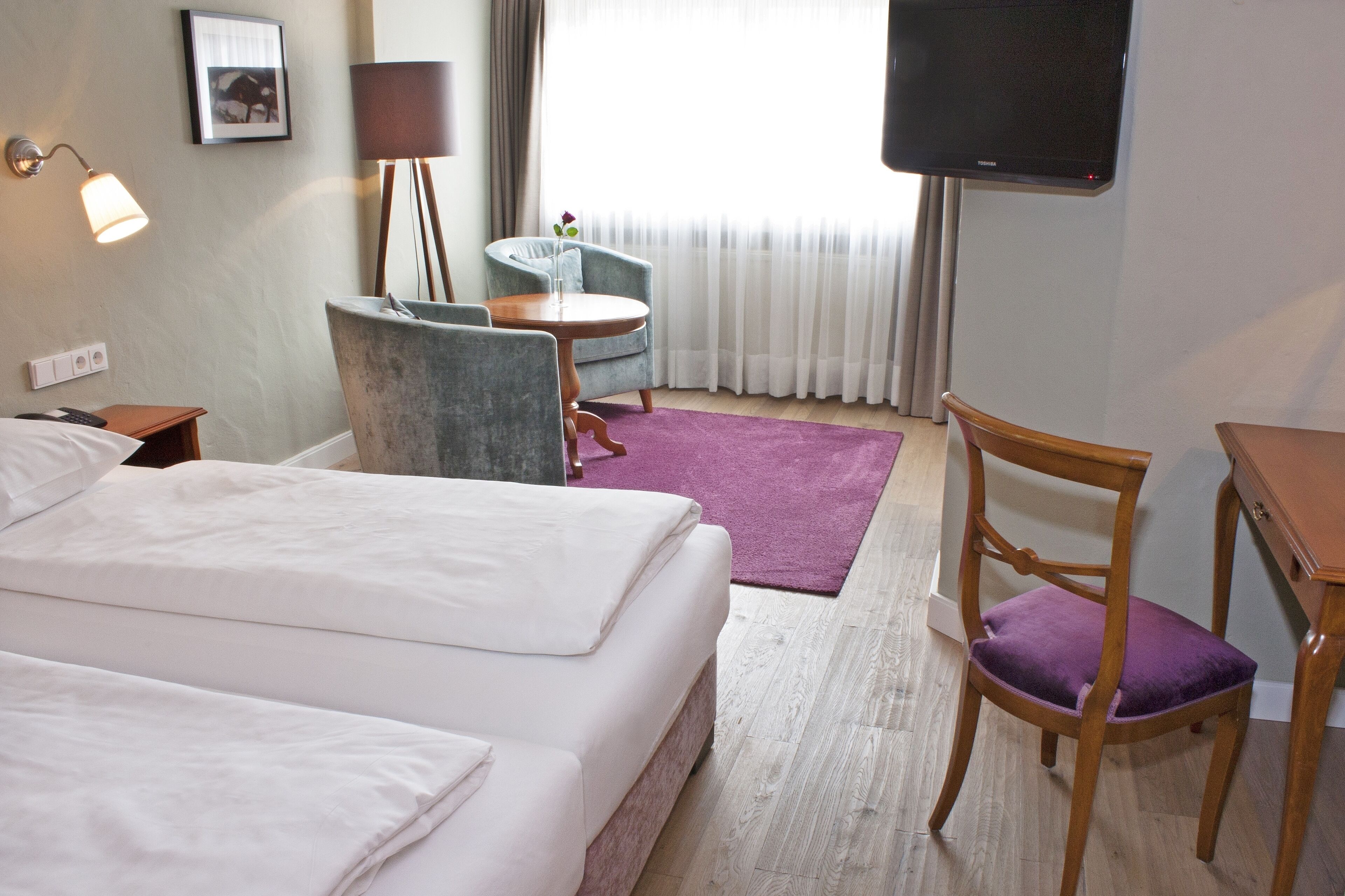 Quarto de hotel com duas camas, uma escrivaninha, uma cadeira, uma TV e um abajur.