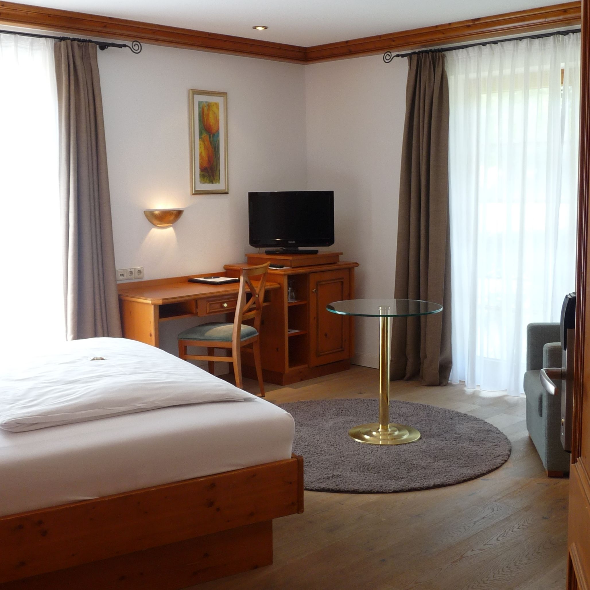 A hotel room with a bed, a desk with a television, a chair, a small table, and a painting on the wall.