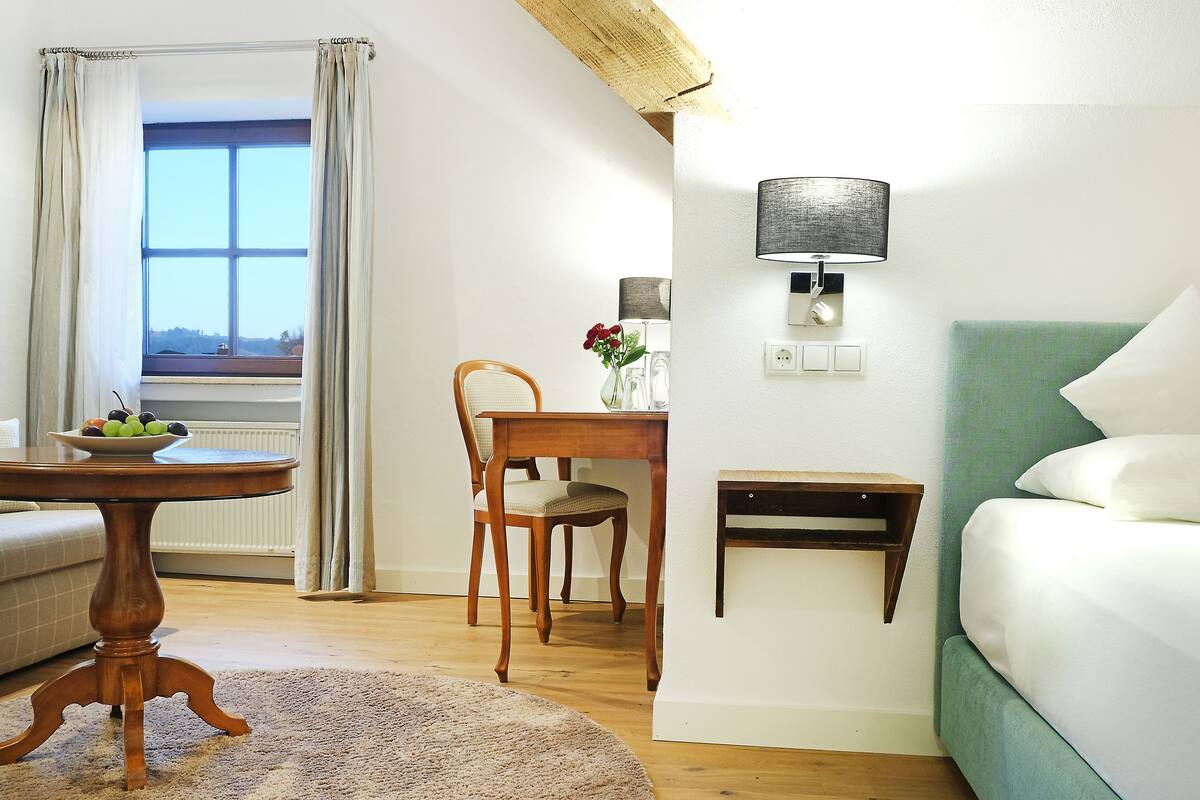 A room with a bed, a table with a bowl of fruit, a chair, a lamp, and a view of the outdoors through a window.