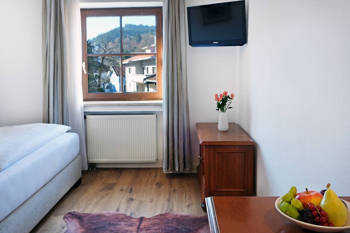 A bedroom with a bed, a wooden dresser, a television, a window with curtains, and a fruit bowl on the dresser.