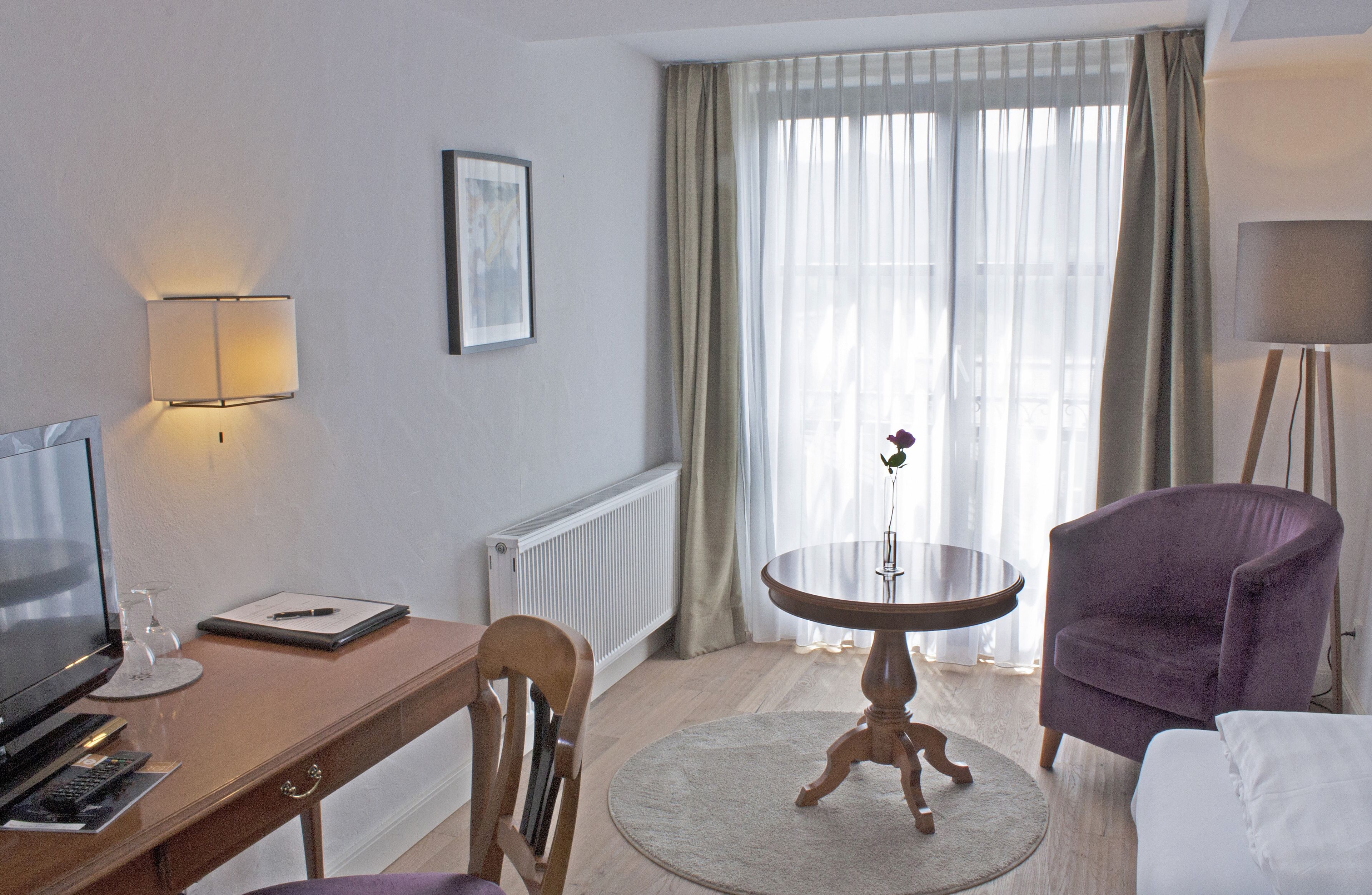 A hotel room with a wooden desk, a television, a round table with a vase, a purple armchair, and a view of the outdoors through sheer curtains.