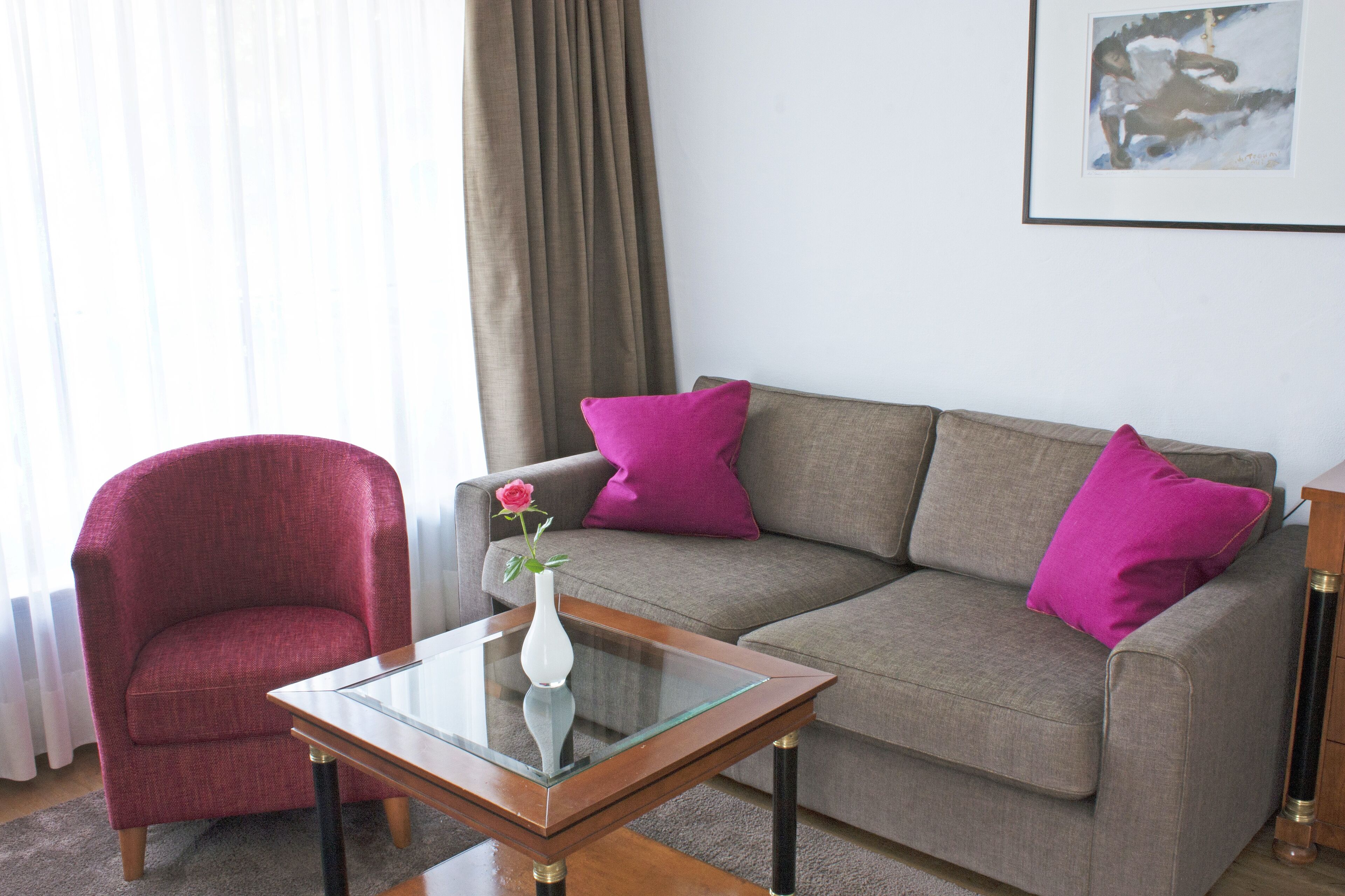 Ein Wohnzimmer mit grauem Sofa, einem roten Sessel, einem Glastisch und einer Vase mit einer Blume.