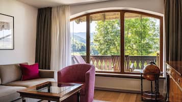 A cozy living room with a sofa, armchair, and coffee table, a large window with a view of greenery and mountains, and a wooden cabinet.