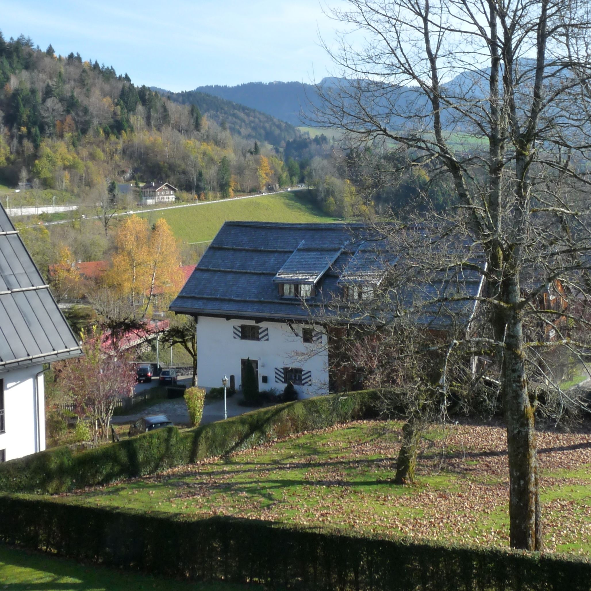 A white building with a dark roof, surrounded by a well-maintained garden and a view of a green valley with trees and hills in the background.