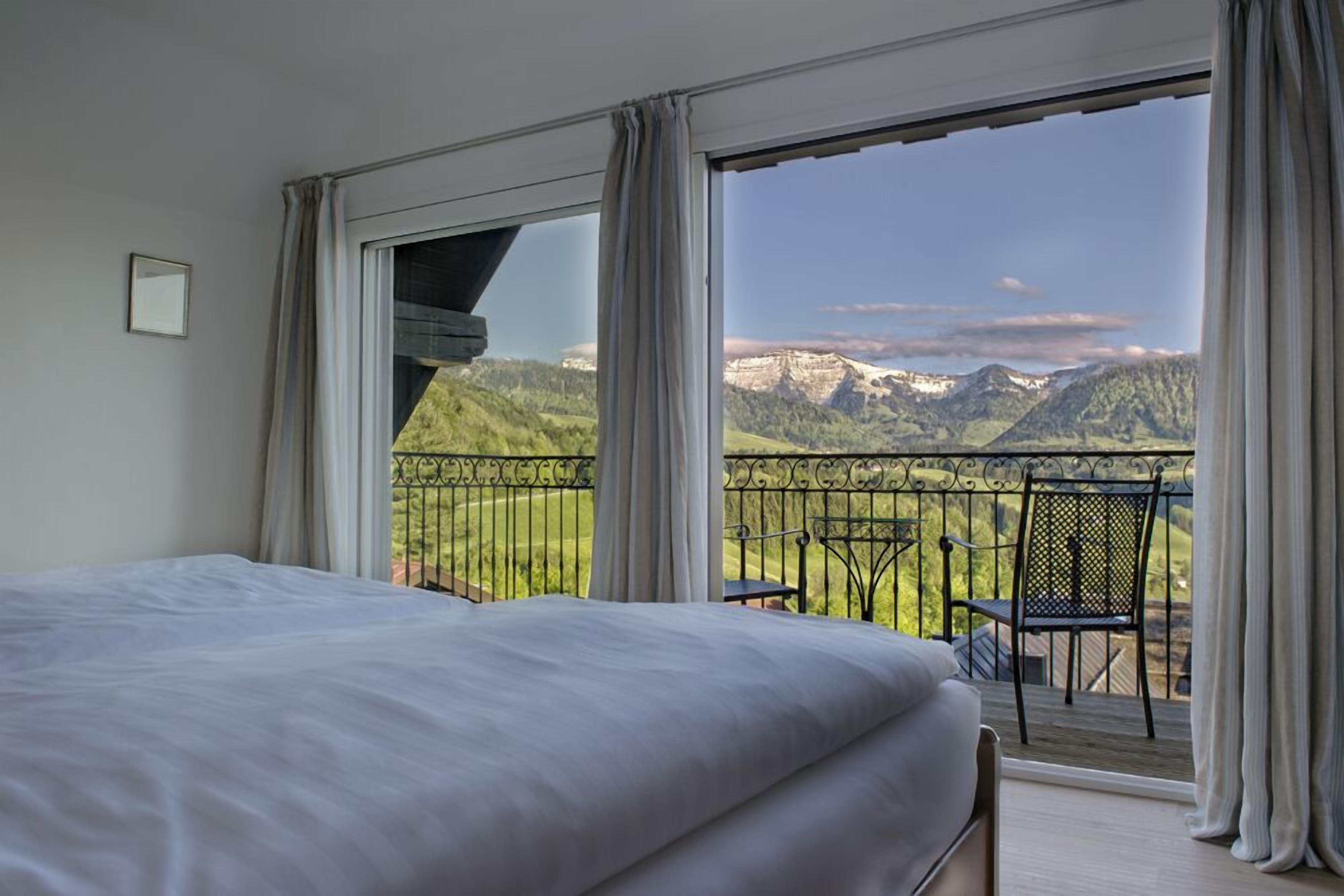 A bedroom with a large bed, a balcony with a view of mountains, and a chair.