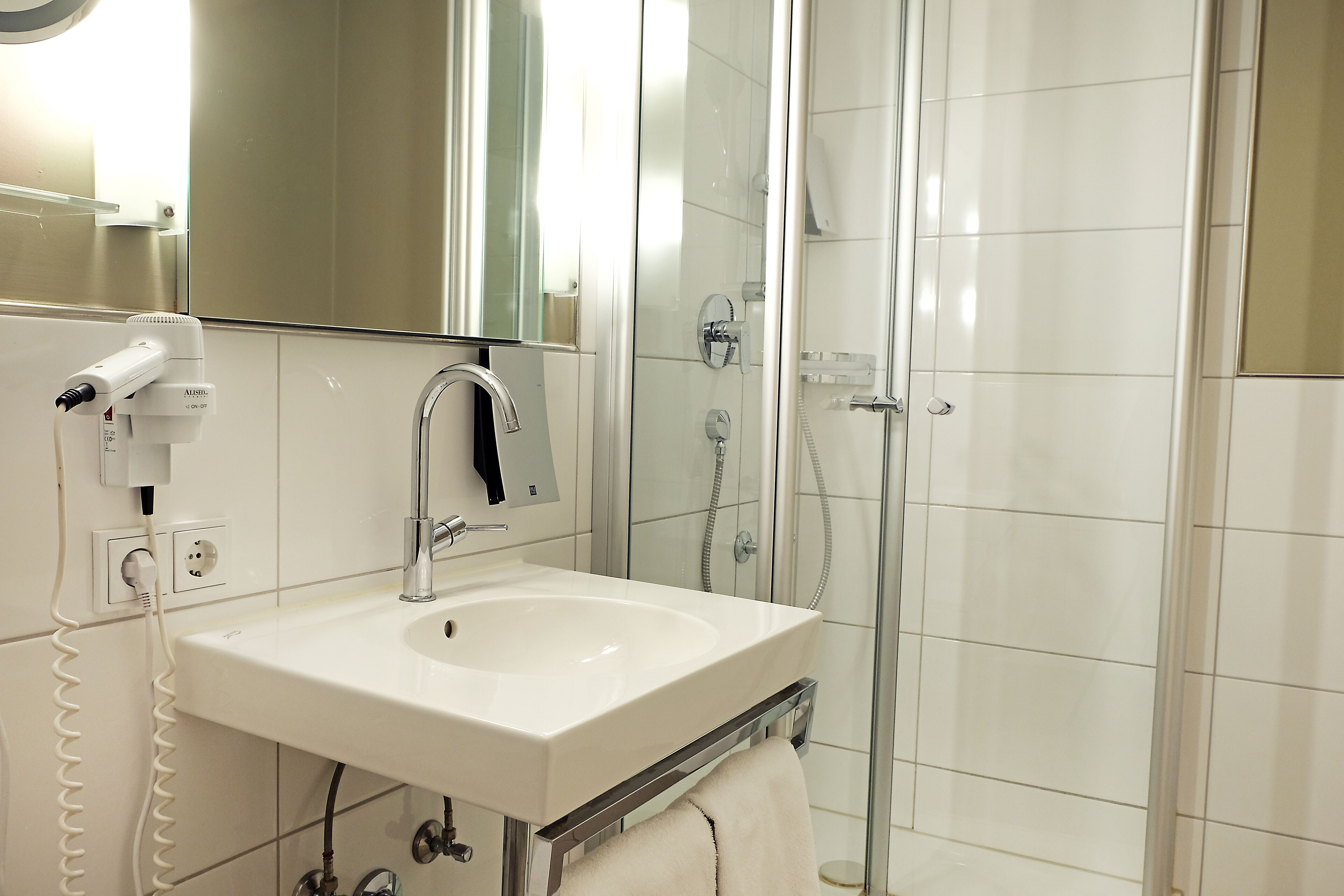 Un baño con lavamanos blanco, espejo, secador de pelo y cabina de ducha de vidrio.