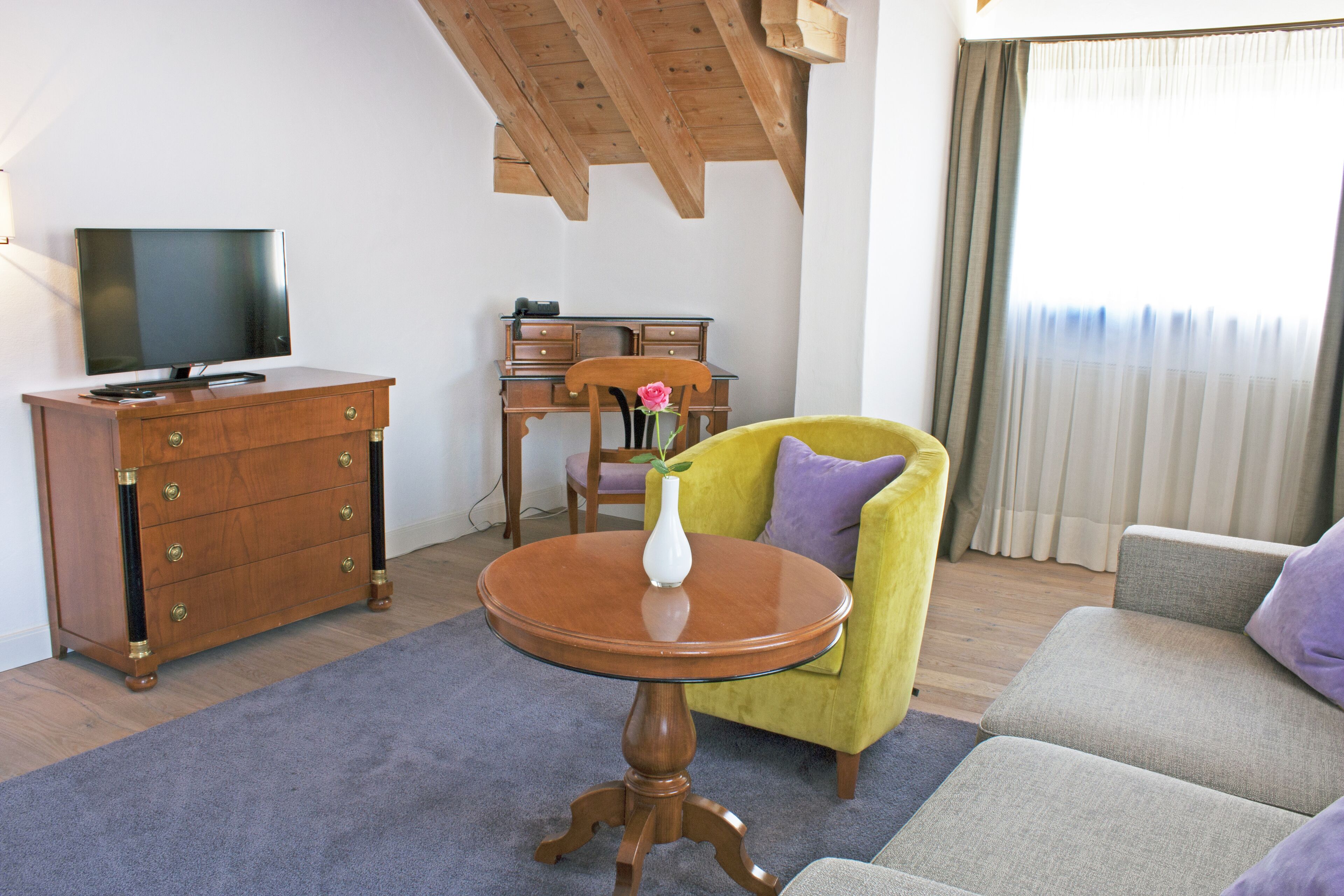 Ein Wohnzimmer mit einem hölzernen Schminktisch, einem Fernseher, einem runden Holztisch mit einer Vase, einem gelben Sessel und einem grauen Sofa.
