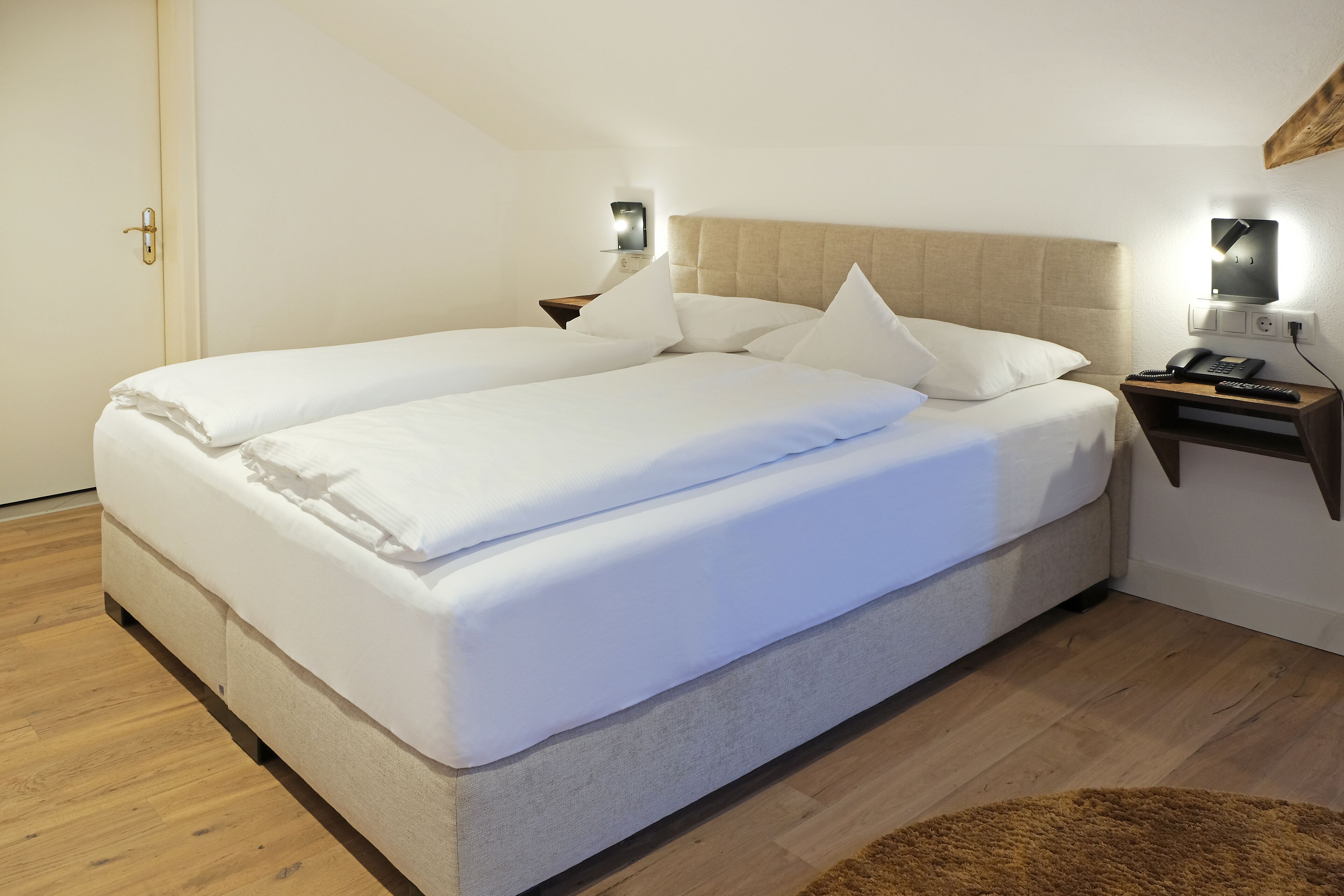 Ein ordentlich bezogenes Doppelbett mit Kopfteil, ein Nachttisch mit Lampe und ein an der Wand befestigtes Telefon.
