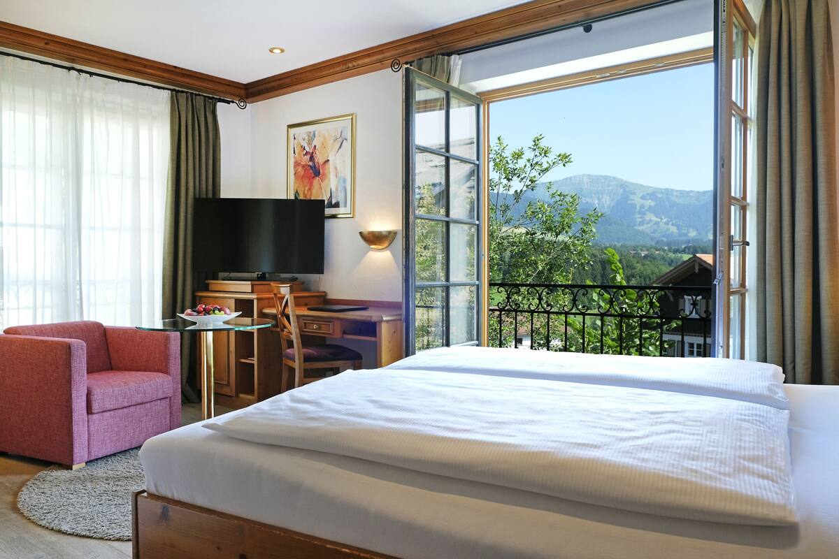 A hotel room with a large bed, a desk with a TV, a chair, and a view of mountains through a large window.