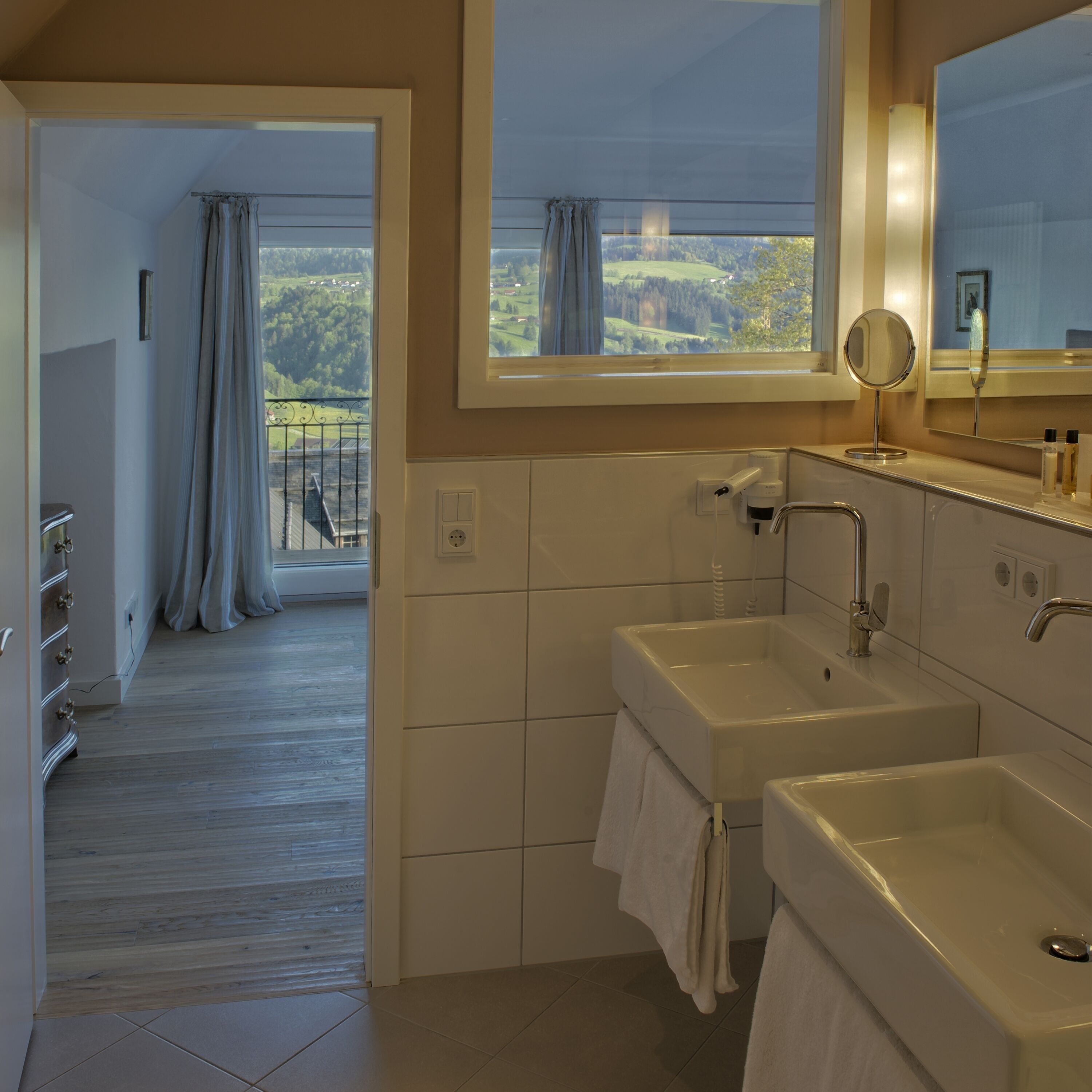 A bathroom with two sinks, a mirror, and a window with a view of a green landscape.
